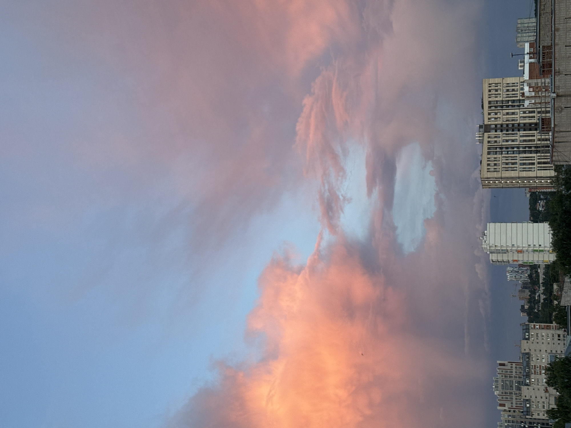 The image shows a dusk cityscape in Izhevsk, Russia. The sky is predominantly blue, transitioning into vibrant peach-colored clouds. On the right side of the frame, various buildings are visible, ranging from high-rise apartments to smaller structures surrounded by trees. The buildings exhibit a mix of architectural styles and colors, including white, gray, and brick. The overall scene captures the juxtaposition of urban development and natural beauty during the evening.