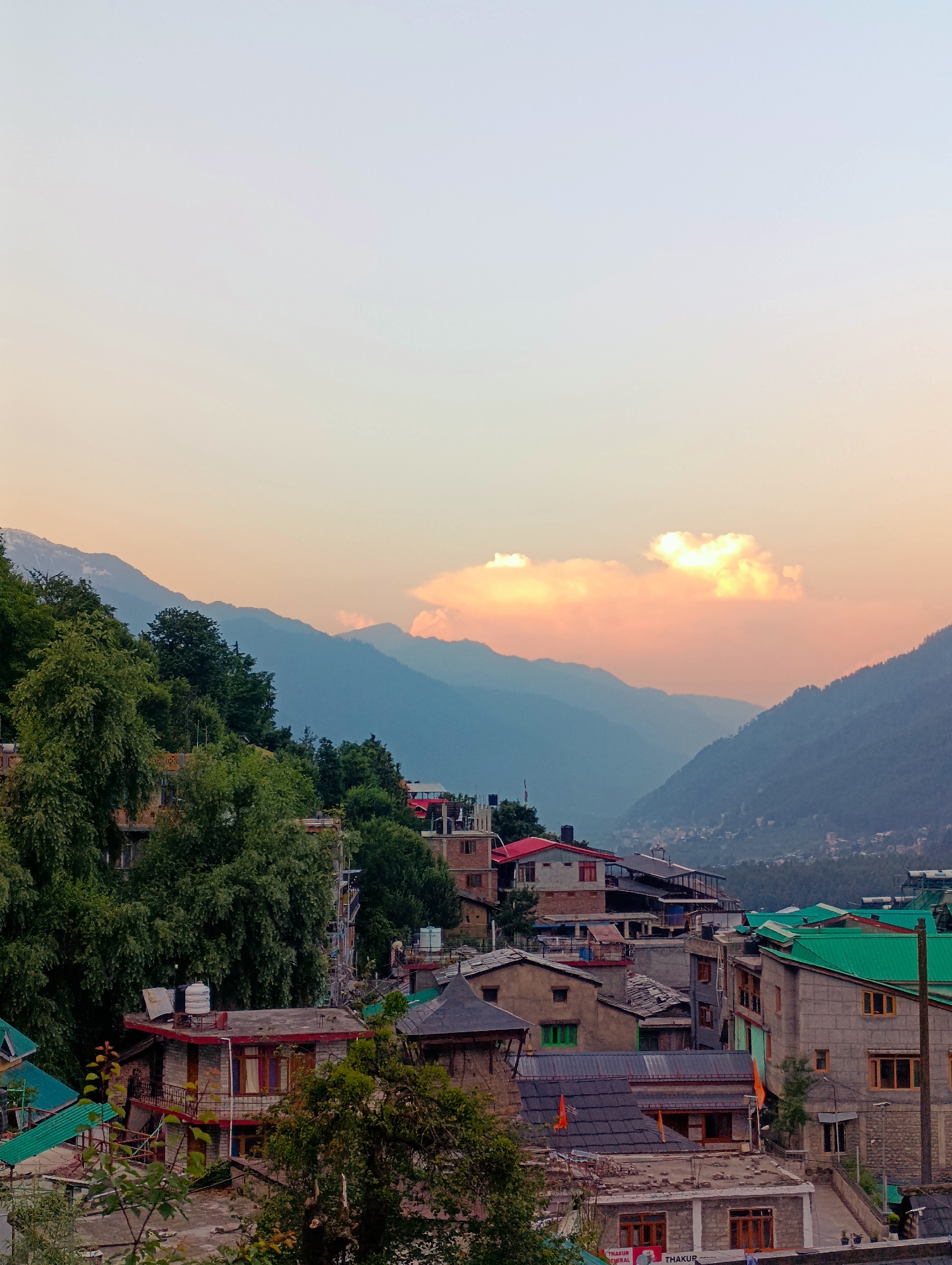 An elevated, vertical, medium shot captures the village of Vashisht, India, nestled within a valley during a sunset. The sky transitions from a pale, soft white at the top to a warm, peachy orange and pink closer to the horizon. A cluster of illuminated clouds is visible in the upper right quadrant, catching the sunset's glow. Beyond the clouds, a series of layered mountain ranges recede into the distance, their forms a deep blue against the lighter sky. The slopes are covered in a dense, green forest.
In the foreground and midground, the village itself is densely packed with buildings constructed from stone and brick. Many structures feature pitched roofs, some covered in dark grey tiles and others in vibrant green metal. Terraced levels and balconies are visible on some buildings. Lush green trees, some quite large and mature, are interspersed throughout the village, partially obscuring some of the structures. Hints of a red truck can be seen in the distance amongst the buildings. Solar water heaters are visible on some rooftops. The overall impression is of a vibrant, lived-in community situated in a dramatic natural landscape.