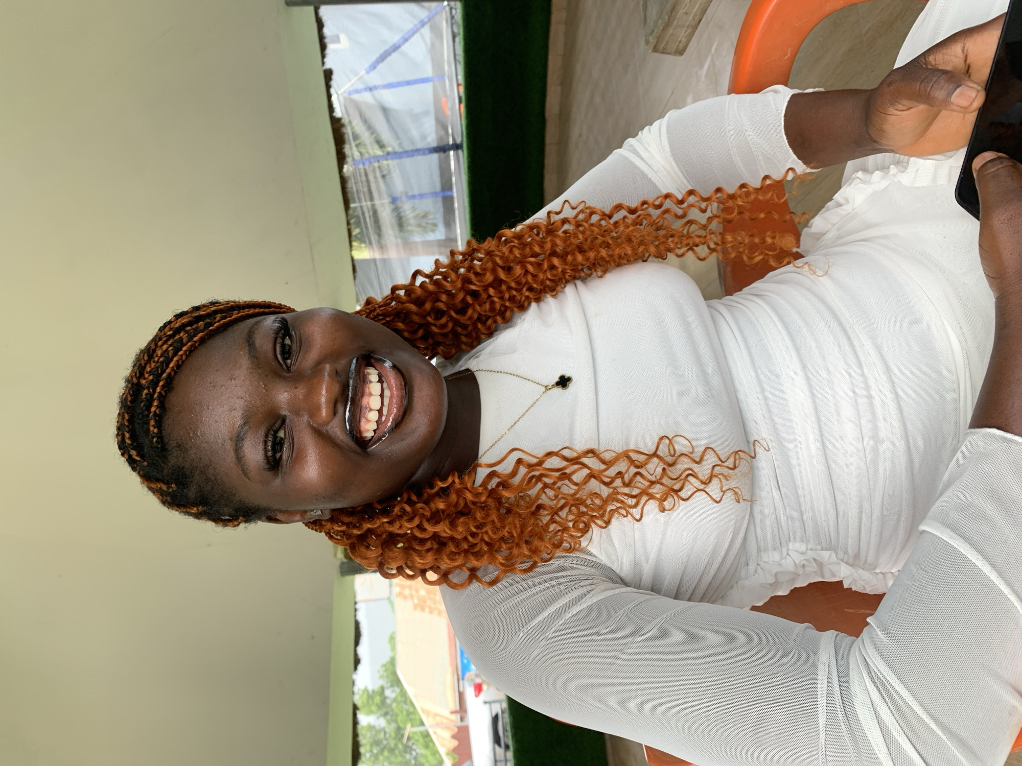 A dark-skinned woman with brown and black braided hair sits on an orange chair. She wears a white dress and a gold necklace with a dark pendant. She is smiling, displaying her teeth, and holding a black smartphone in her right hand. The background shows a glimpse of the outdoors with green vegetation and a semi-transparent structure. The location is in Adankolo, Nigeria.