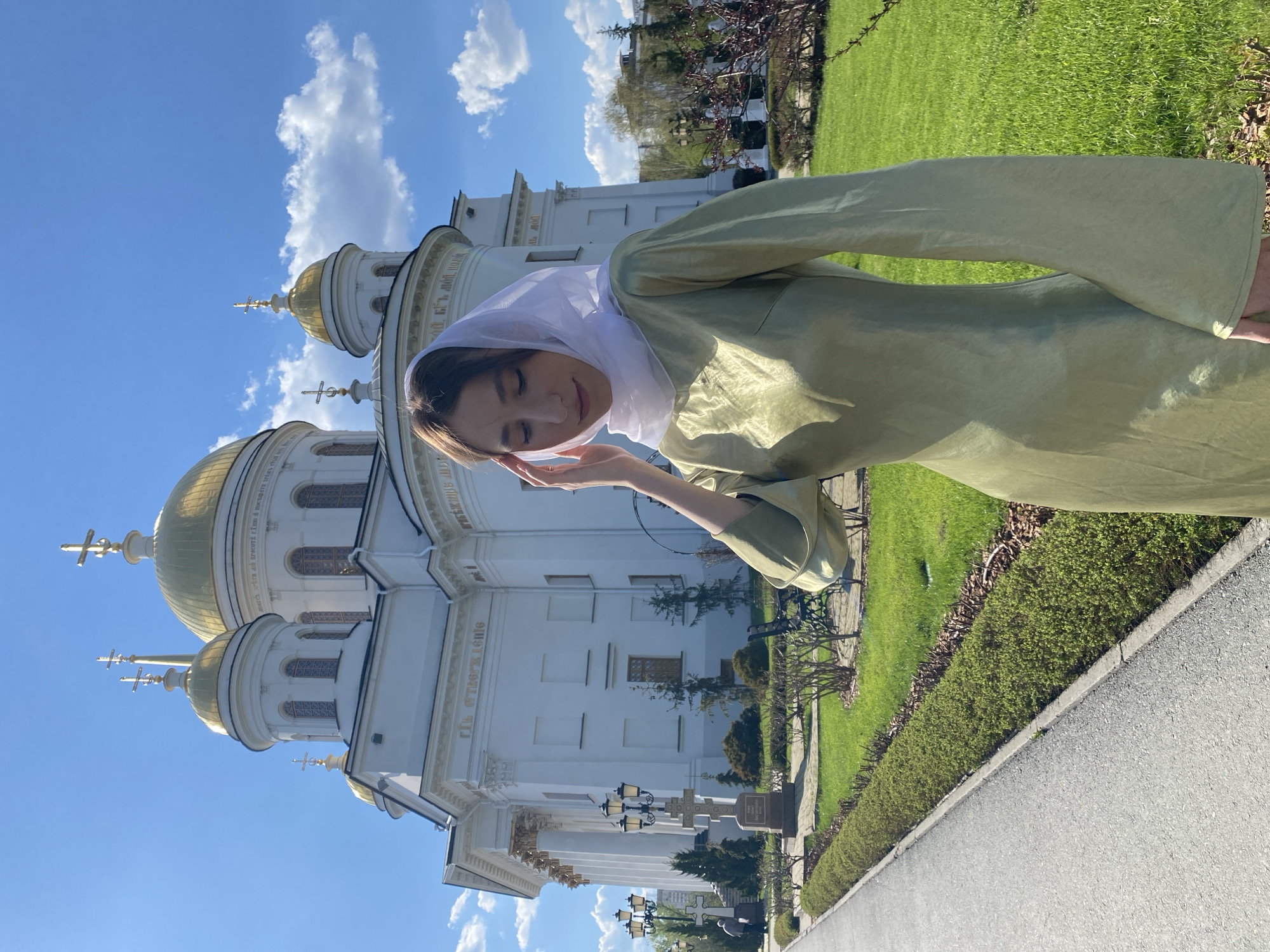 A woman in a light green garment and white headscarf stands in front of the Church on the Blood in Yekaterinburg, Russia. The church, a large white building with gold domes, is visible in the background against a blue sky with white clouds. The woman holds her hand near her face with her eyes closed. The church sits on the left side of the image, while green grass and a paved walkway are visible on the right. Street lamps line the walkway.