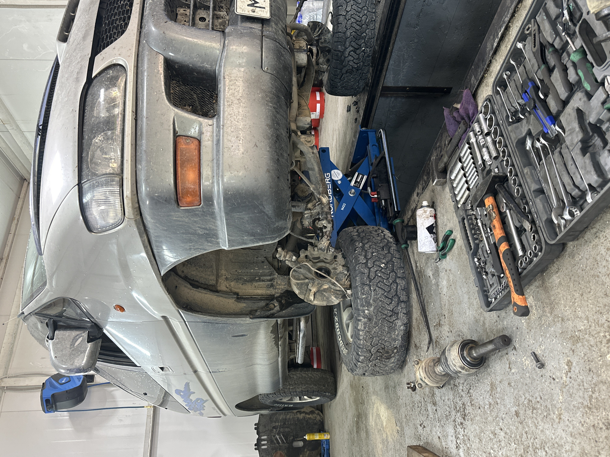 In Blagoveshchensk, Russia, a light-colored van is undergoing repair work in a garage. The vehicle is elevated on a blue hydraulic jack, with its front wheel removed and resting on the floor. A disassembled drive shaft lies on the ground next to the van. A toolbox filled with various tools, including wrenches, sockets, and a hammer, is also visible on the floor. The garage environment appears to be industrial, with concrete flooring and white walls. The scene suggests an ongoing automotive repair operation.