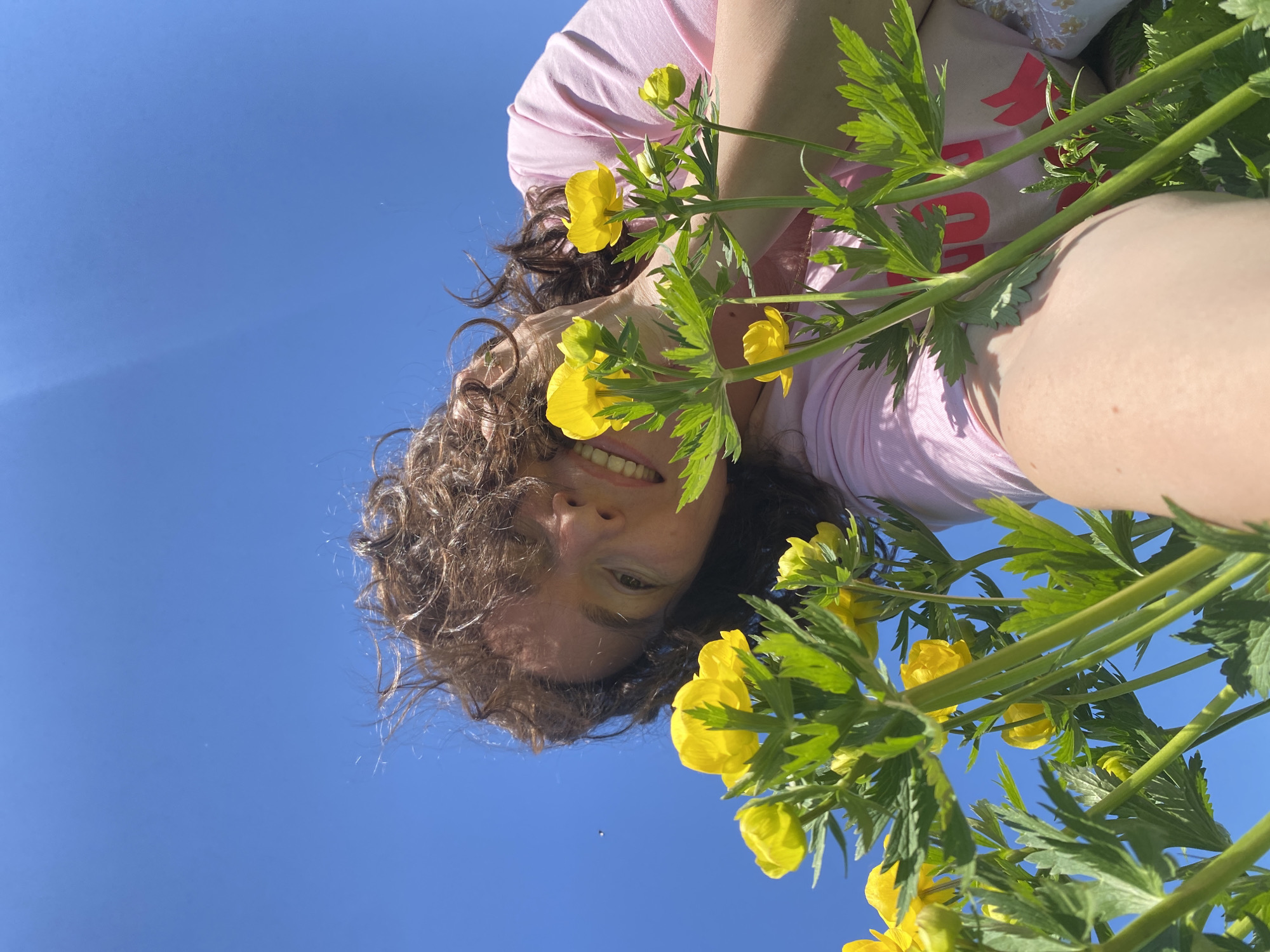 A low-angle, slightly upside-down shot shows a person with curly brown hair and a pink t-shirt holding yellow globe flower stalks against a clear blue sky. The person's face, with a visible smile, is partially framed by the yellow flowers and green foliage. The location is not discernible from the image.