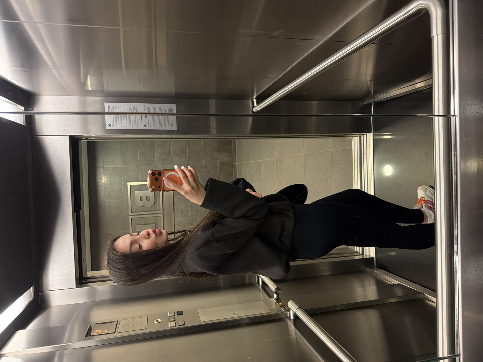 A woman is taking a selfie in an elevator. She is holding a phone in her right hand, visible due to its orange phone case. The woman is wearing a black jacket, black leggings, and white sneakers with orange accents. She has long, dark hair. The elevator has stainless steel walls and handrails. There is a control panel with buttons and a display indicating the floor level. A logo, partially obscured, is visible on the wall behind the woman. Based on the logo, it is likely that the image was taken in the elevator of the B1 restaurant in Sofia, Bulgaria.