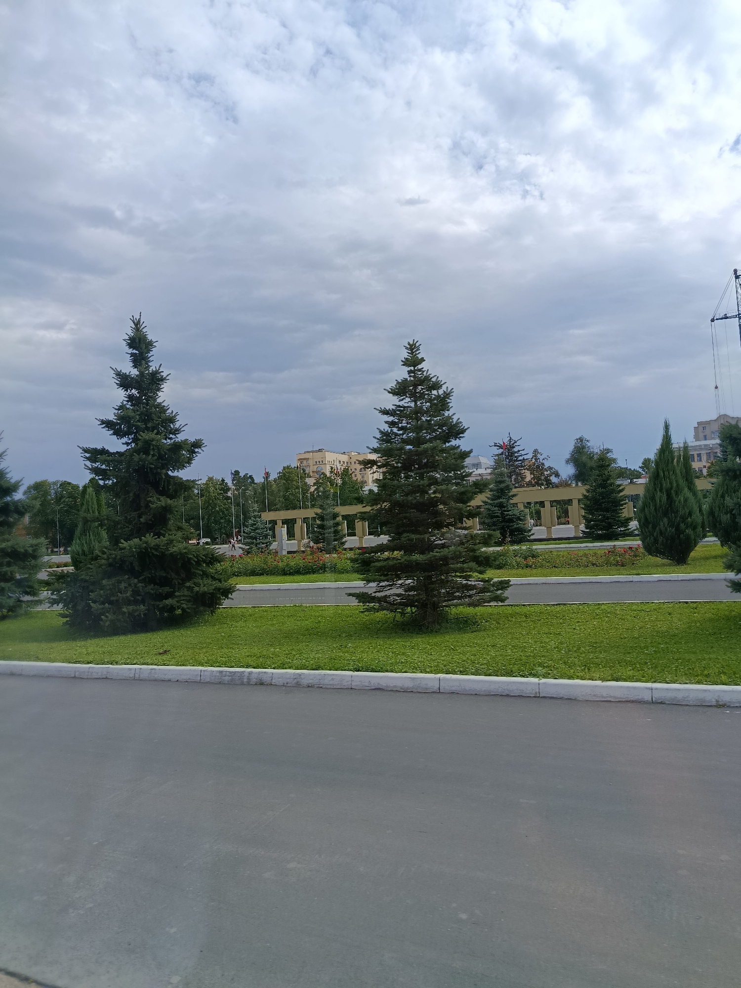 A wide-angle, eye-level outdoor shot shows a paved road in the foreground, transitioning to a well-maintained grassy area with several evergreen trees. Behind the trees, a row of white pillars supporting an arched structure suggests a park or memorial. In the distance, a multi-story building with a light-colored facade and darker roof is visible, along with some smaller trees and what appears to be a construction crane on the far right. The sky is overcast with numerous clouds. The scene is likely located in Saratov, Russia, given the architectural style and vegetation. No people are clearly visible.