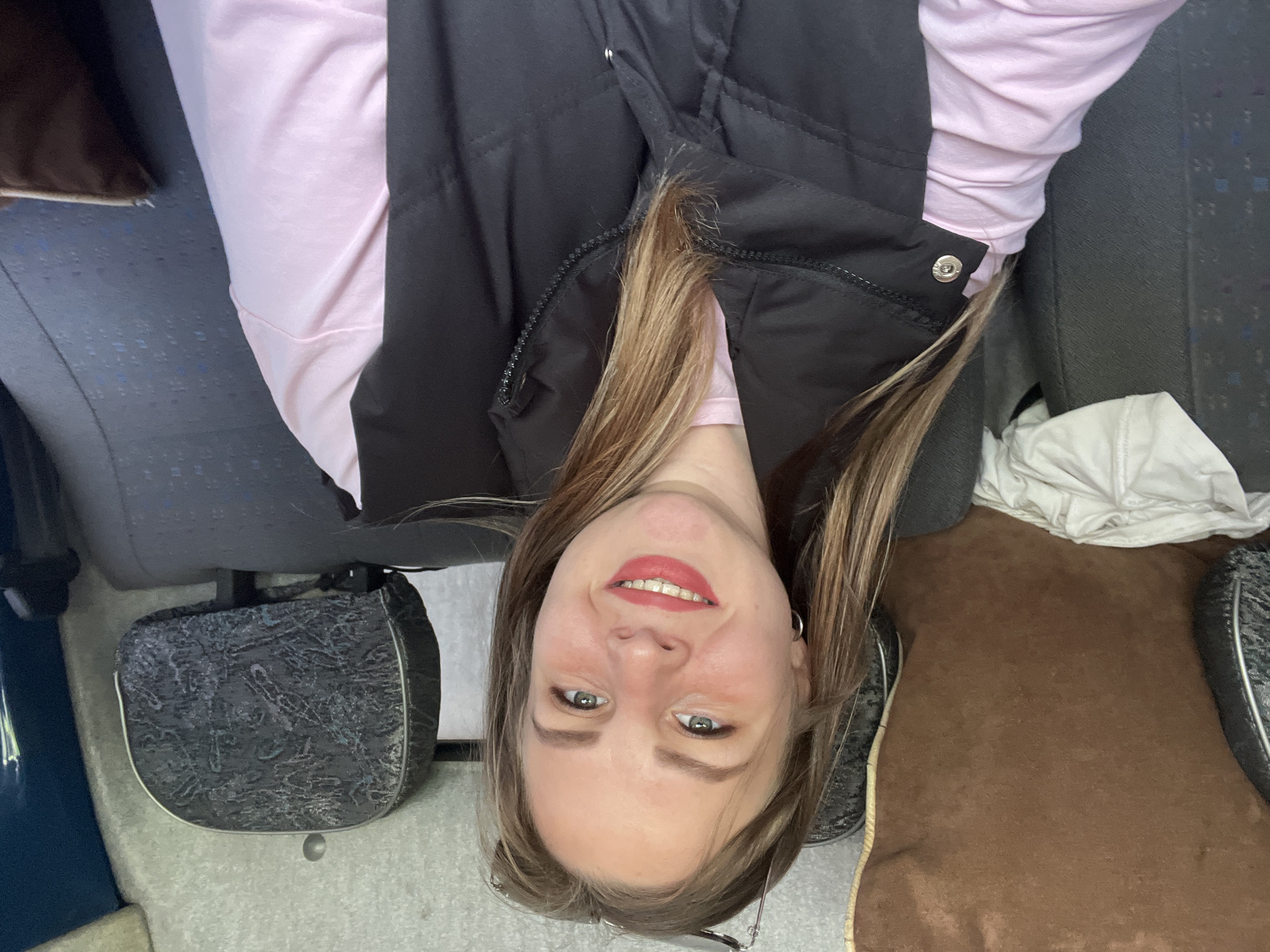 A woman with long brown hair, wearing a pink shirt and a black vest, is sitting in what appears to be a vehicle. The background shows patterned seats, potentially in a bus or van. Details to confirm the location in Izhevsk, Russia, are absent.