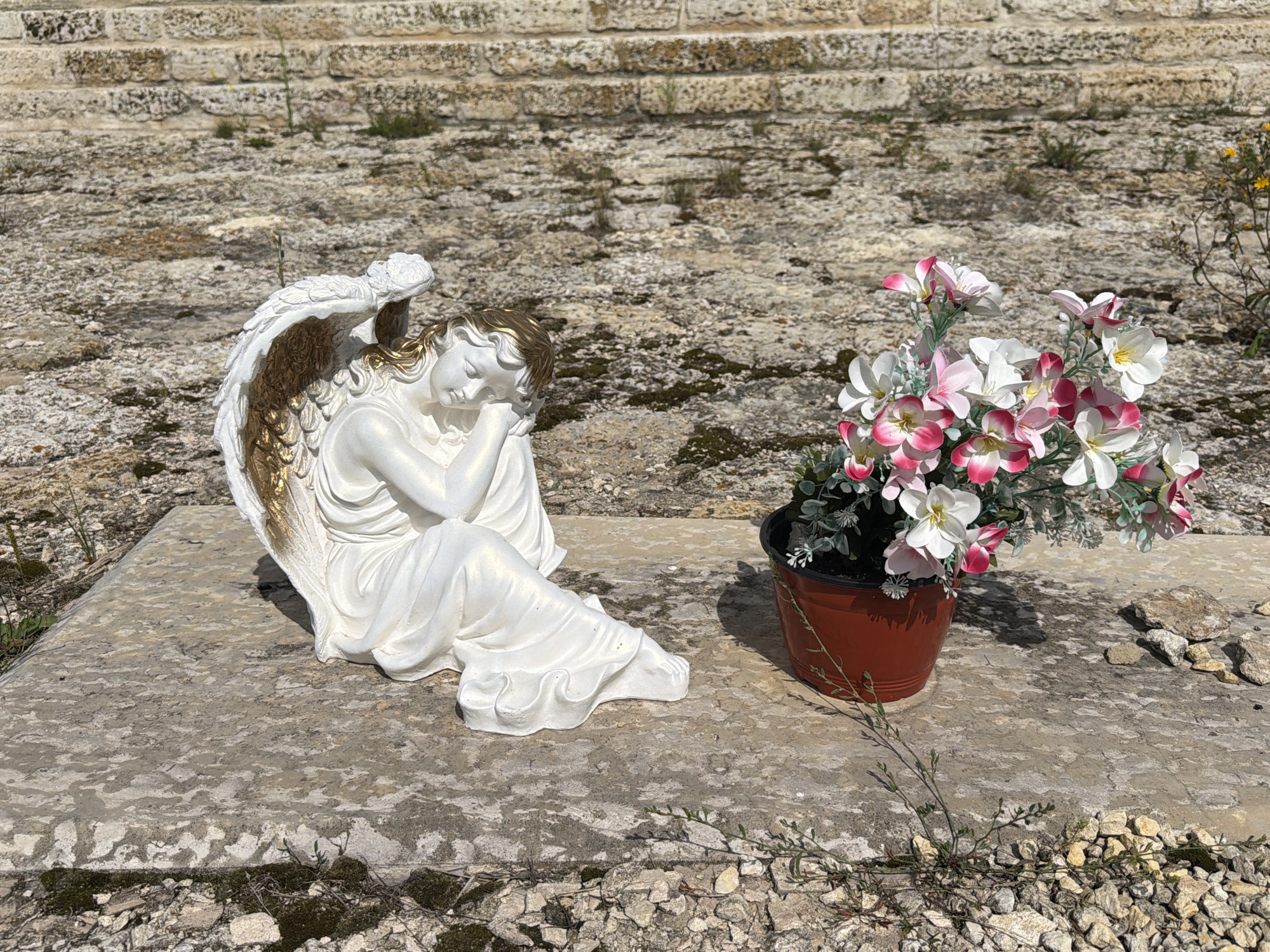 An angel statue, made of white material with gold accents on its wings and hair, is positioned on a flat, weathered concrete slab. The statue depicts the angel in a seated position, with its head resting on its arm, appearing to be asleep or in deep thought. To the right of the statue, a brown pot contains a bouquet of artificial flowers in various shades of pink and white. The slab is set on a ground covered with gravel and small stones, with patches of dry moss and sparse vegetation. In the background, a textured wall constructed of rough, tan-colored stones forms horizontal bands. The scene is bathed in bright sunlight, casting shadows on the concrete. The overall setting appears to be an outdoor area, possibly a cemetery or memorial site, given the presence of the statue and the nature of the surroundings. The location is identified as Скорняково, Russia.