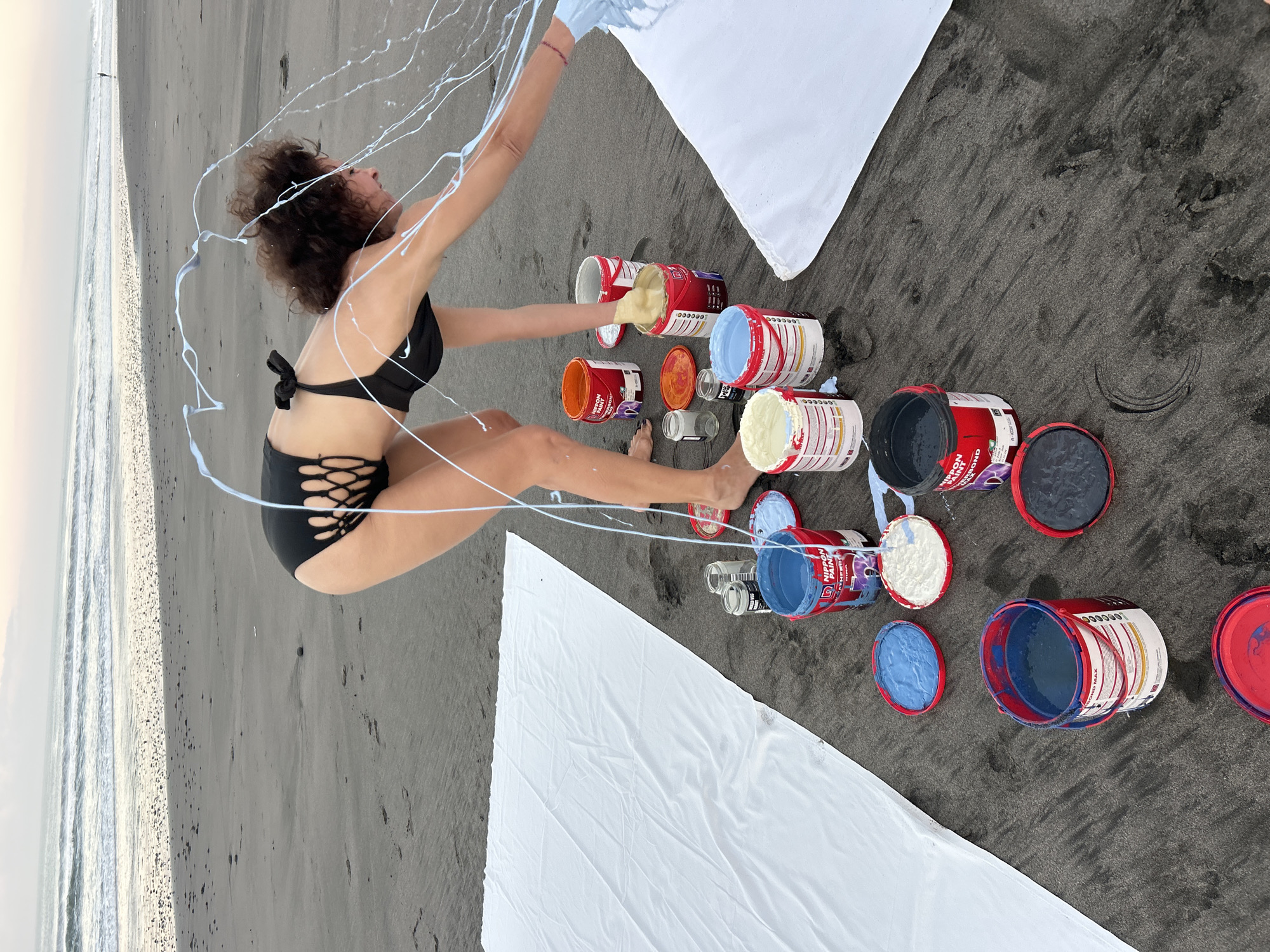 A woman in a black cutout swimsuit is on a dark sand beach, bending over and splashing white paint from a container with her hand. Several open buckets of paint in blue, white, and orange are scattered on the sand around her. Two large white sheets are laid out on the sand, one to the left and one to the right of the scene. The ocean with gentle waves is visible in the background, under a hazy sky. The location is the beach in Tabanan, Indonesia.