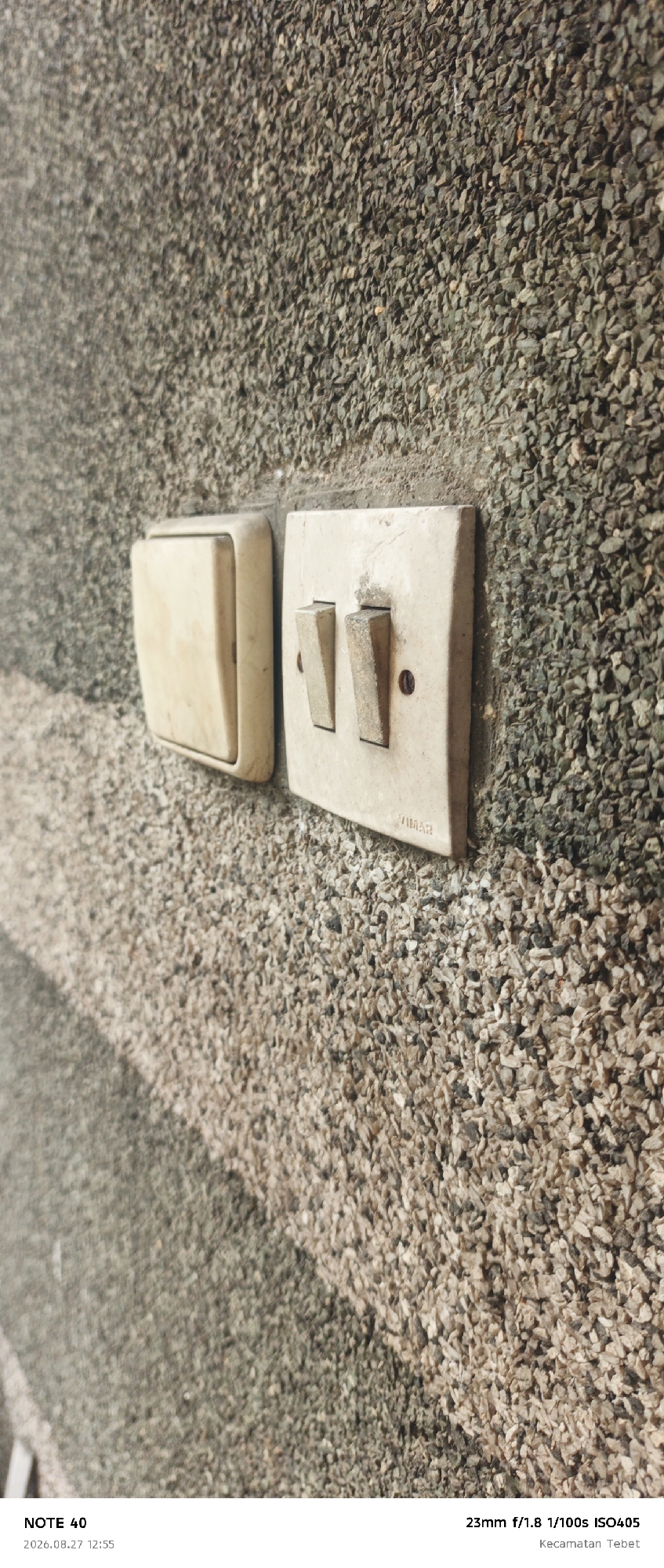 A close-up, eye-level shot shows two light switches on a textured, pebbled wall. The switch on the left is a single, cream-colored toggle switch. To its right is a double toggle switch, also cream-colored, with both toggles in the down position. The wall has a rough, granular texture, with various shades of grey and brown pebbles. A diagonal shadow stripe runs across the lower left portion of the wall, creating a contrast between a lighter and darker textured area. Text at the bottom indicates "NOTE 40", "2026.08.27 12:55", "23mm f/1.8 1/100s ISO405", and "Kecamatan Tebet". This suggests the image was taken in Tebet, South Jakarta, Indonesia.