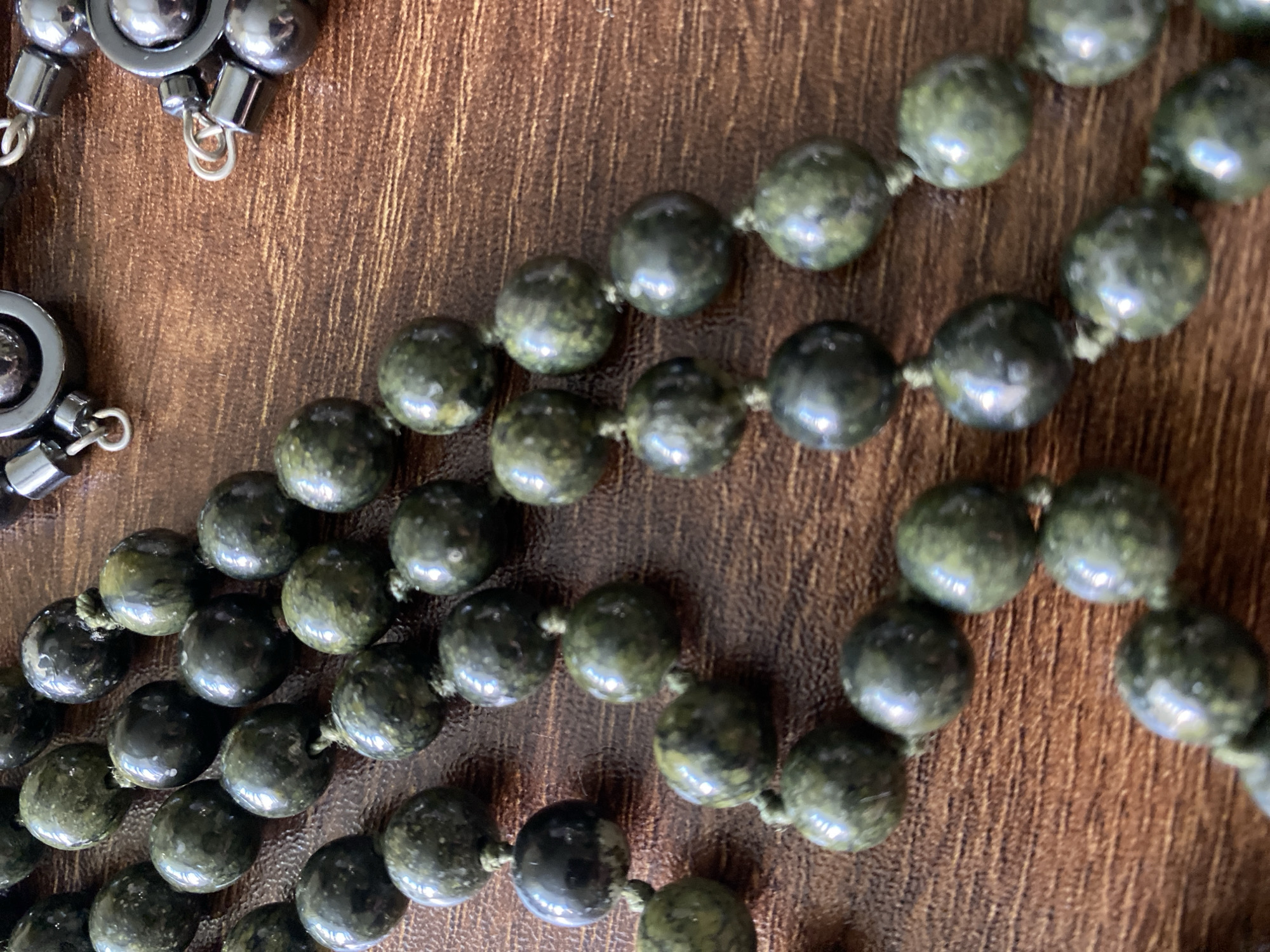 A close-up, overhead shot shows several strands of beaded jewelry on a wooden surface. The beads are mostly round and appear to be made of a dark green, mottled stone, possibly jade or serpentine. The beads are threaded onto a light-colored string, with visible knots between each bead. Some of the beads have a glossy sheen, reflecting the light. In the upper right portion of the image, there are two dark, metallic clasps or connectors, each with smaller metallic rings attached, suggesting the jewelry is a necklace or bracelet. The wooden surface has a visible grain pattern and a warm, brown hue. The lighting is direct, creating highlights and shadows on the beads and the wood.