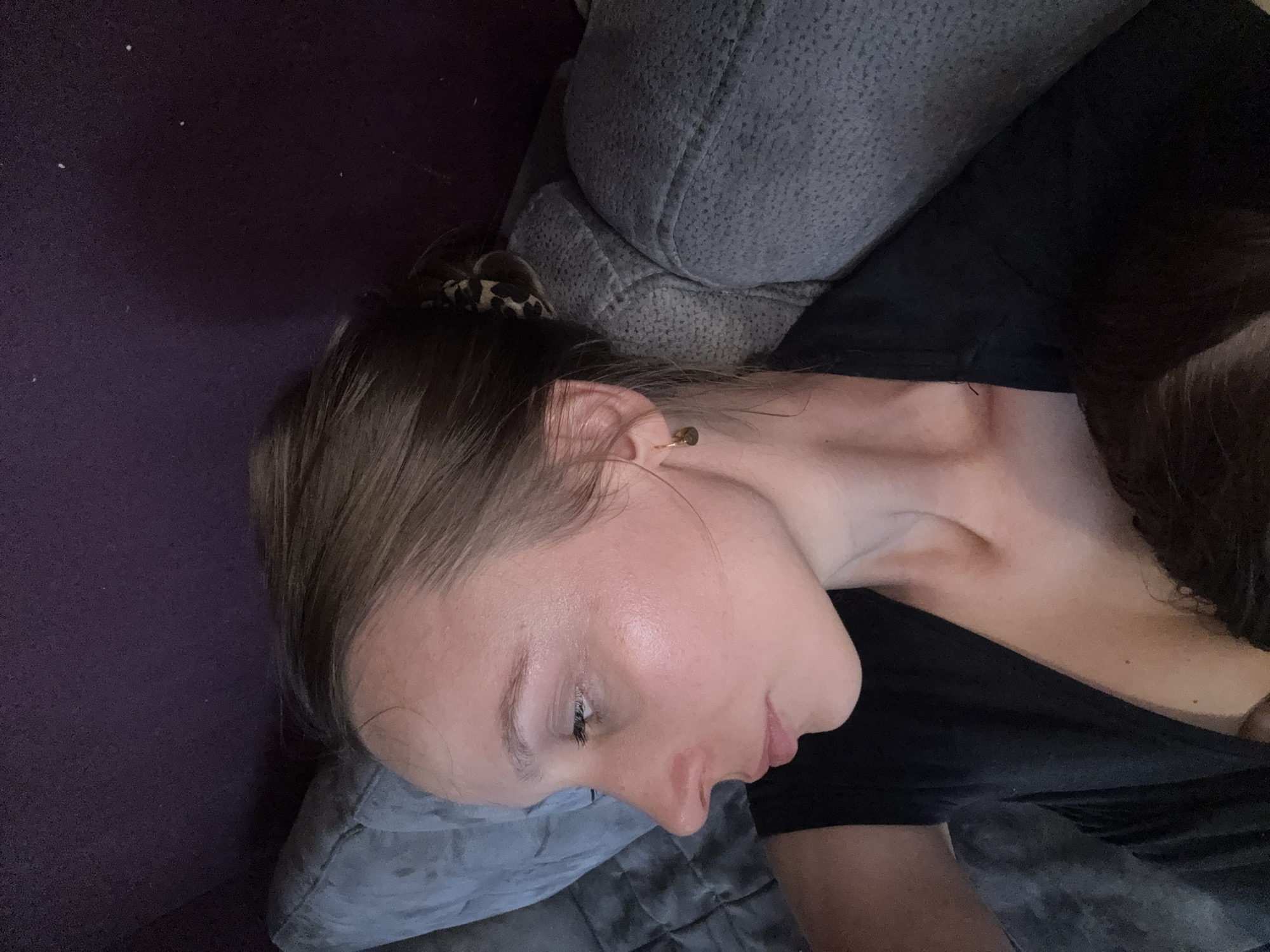 The image depicts a close-up, profile view of a young Caucasian woman lying down, possibly on a sofa, with a dark purple background and a gray textured surface to her left. Her hair is pulled back in a bun with a leopard-print scrunchie. She is wearing a black t-shirt. She has light skin with some visible highlights on her cheekbones and forehead, suggesting makeup or natural shine. A small, gold hoop earring is visible in her left ear. The lighting appears to be artificial and somewhat dim. There are no discernible elements that indicate a specific location within Überherrn, Germany. The image focuses tightly on the woman's face and upper body, with the background elements blurred and out of focus.
