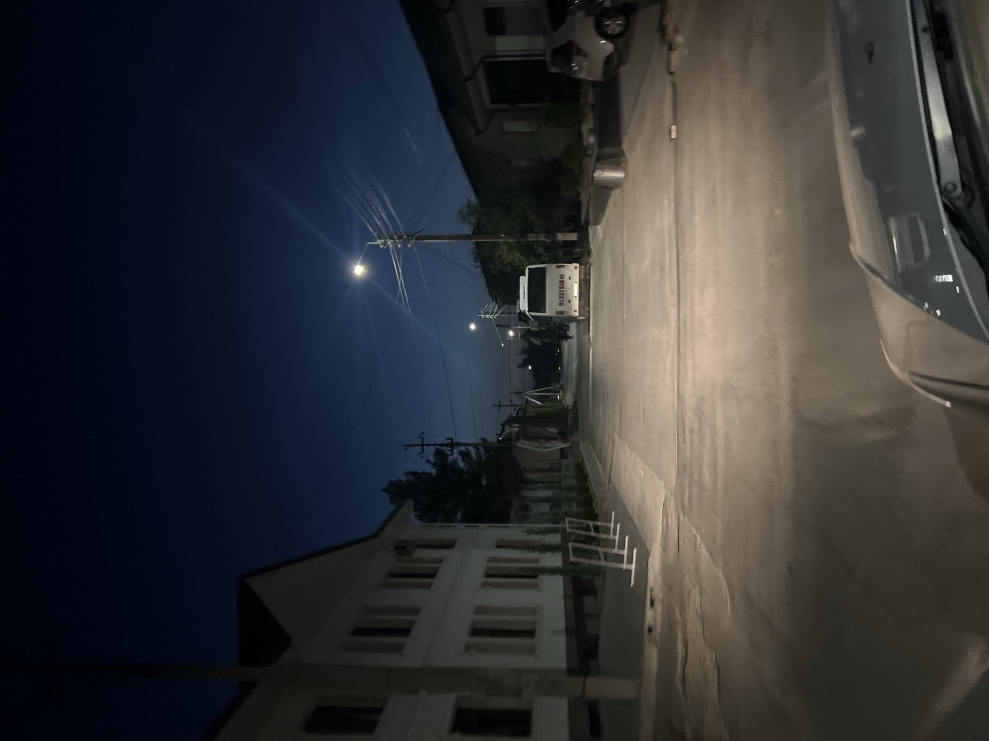 The image shows a street scene at dusk or night in Tashkent, Uzbekistan. A white bus with the license plate "01 337 SIA 07" is parked in the middle of the street, facing away from the camera. To the left of the bus, a white building with multiple windows is visible. On the right side of the street, a white car and a grey car are parked. Streetlights illuminate the scene, casting long shadows. The sky is a deep blue, characteristic of twilight. The road appears to be paved. The image is taken from the perspective of a vehicle driving down the street.