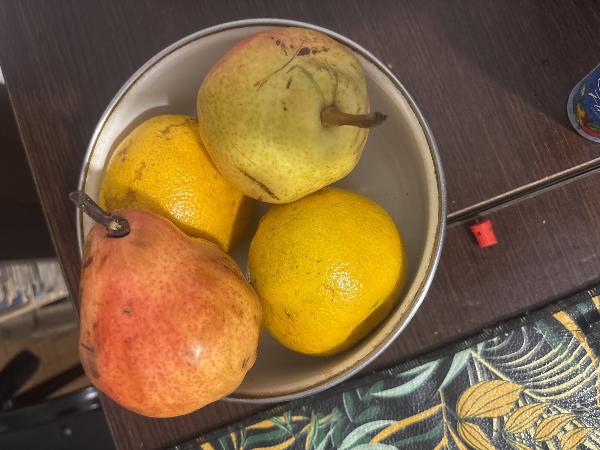 A top-down shot shows a bowl containing two pears and two citrus fruits, possibly oranges. The bowl, a light tan color with a darker rim, rests on a dark brown surface, possibly a table or countertop. A patterned surface, possibly a placemat with a dark background and botanical design, is visible near the bottom edge of the frame. A small red object is located adjacent to the edge of the bowl on the dark brown surface. The scene appears to be indoors. Location information about Buenos Aires, Argentina, is not discernible from the image.