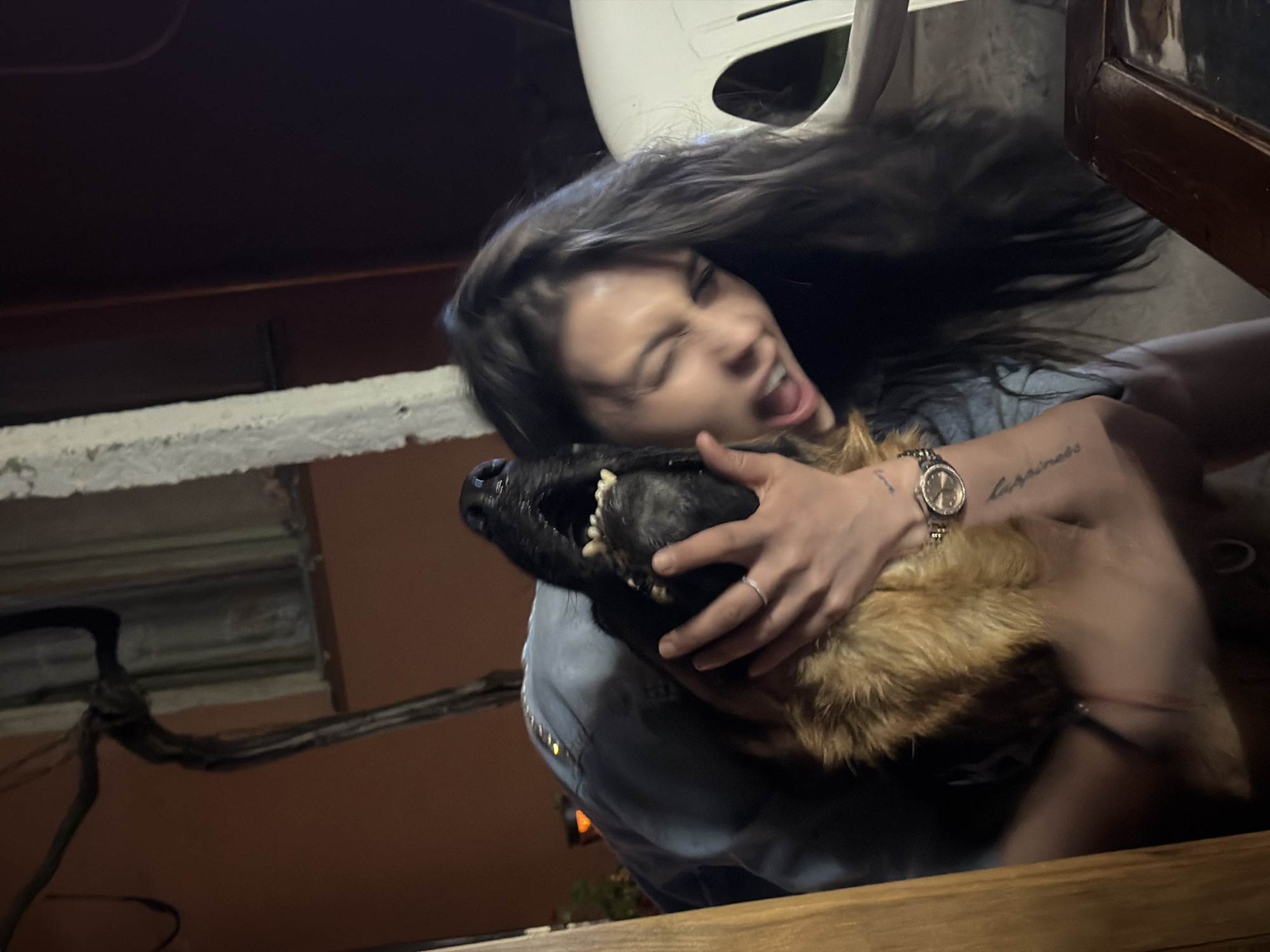 The image shows a woman and a dog in what appears to be an outdoor setting at night. The woman, with long dark hair, is holding the dog close, with her eyes closed and mouth open in an exaggerated expression. The dog, a German Shepherd, has its mouth open as well, revealing its teeth. Both the woman and the dog are close to the camera, making them the central focus. The background is dark and somewhat blurry, but suggests an urban environment with buildings and some vegetation. The presence of a wooden table in the foreground and a white chair in the background hints at a patio or outdoor seating area. There is a vine-like structure with dark branches visible on the left side of the image. The overall lighting suggests artificial light sources, possibly from nearby buildings or lamps. The location of Golyam Chardak, Bulgaria is not evident from the visual cues in the image itself.