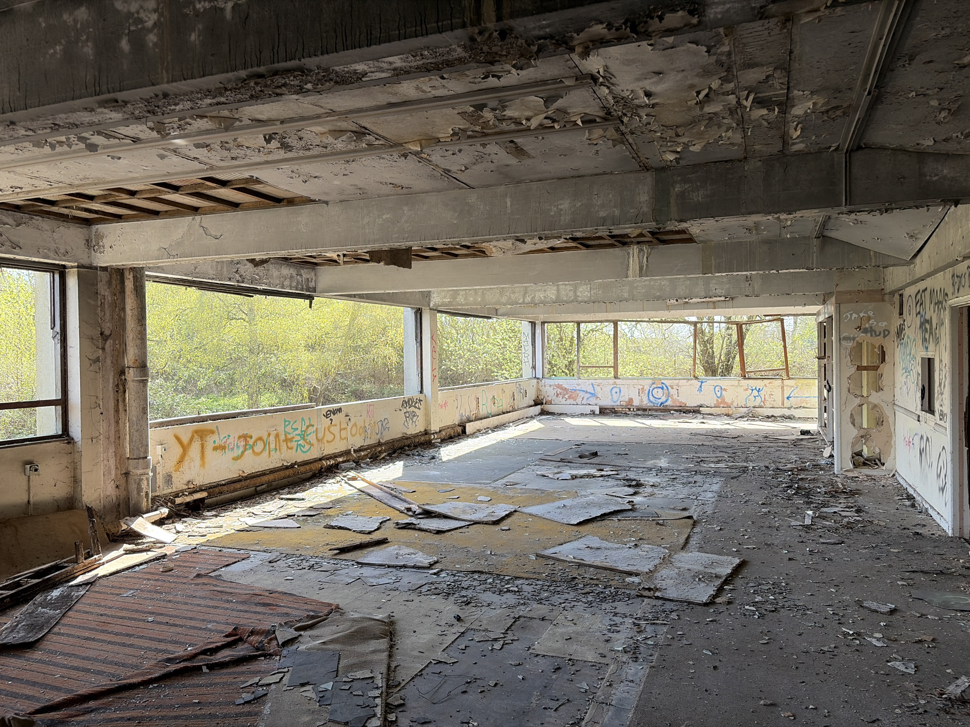The image shows the interior of a derelict building, likely a former office space. The location is consistent with being in Mid Suffolk, United Kingdom, but this is unverified. The room has large windows that provide a view of a wooded area with green foliage. Graffiti is visible on the wall beneath the windows. The ceiling is damaged with exposed structural elements. The floor is covered in debris and remnants of flooring. The room is empty and devoid of people.