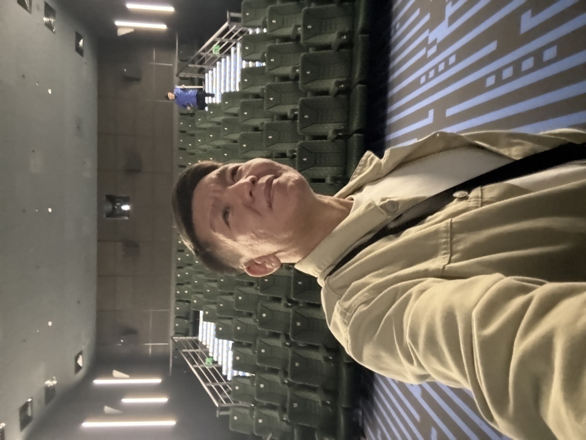 A person is taking a selfie in an empty movie theater. The person is wearing a light-colored jacket over a white t-shirt. The theater has rows of dark green seats, and the floor has a geometric pattern of blue and white lines. In the background, another person is walking up a staircase. The lighting in the theater is dim, with vertical lights on the walls and lights on the stairs.