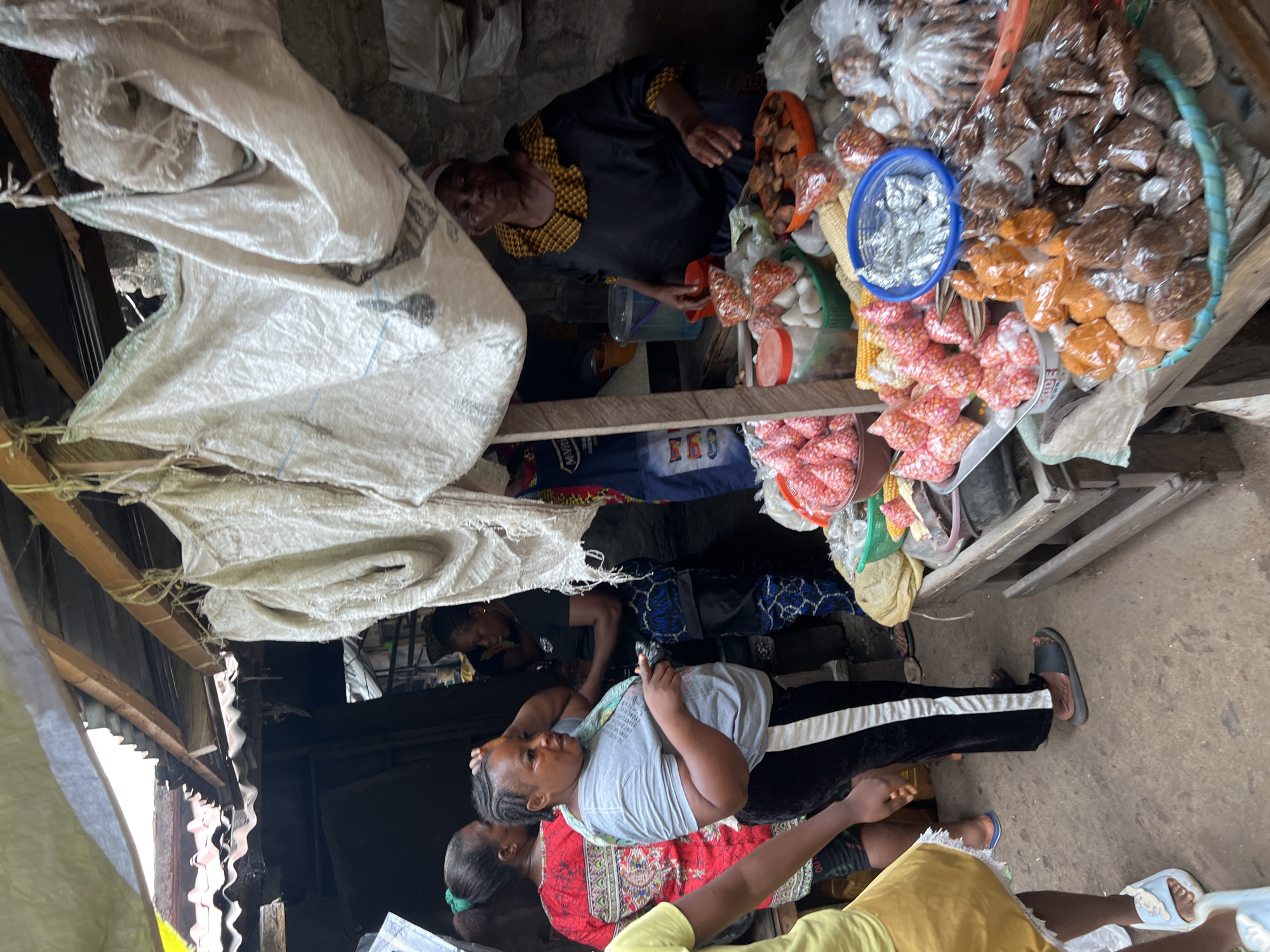 The image depicts an outdoor market scene, with people interacting around a stall laden with various packaged goods. An elderly woman in a black and yellow top is attending the stall. In front of the stall, a woman in a grey shirt and black pants appears to be a customer. There are other people in the vicinity. The stall displays an array of food items packaged in small bags and containers. The environment seems to be a simple structure, possibly with corrugated metal roofing. There are bags, possibly used for storage or transport, visible above the stall. The location is within the city of Unknown, Unknown.