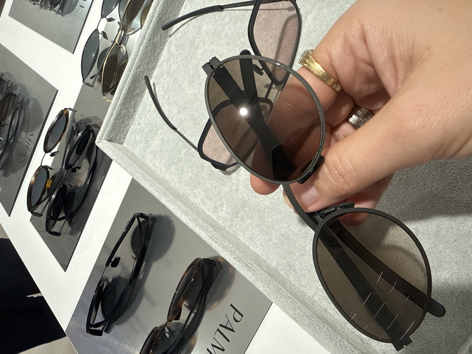 A person's hand, adorned with two rings, holds a pair of dark-rimmed sunglasses with tinted lenses. The sunglasses have thin, dark temples and appear to be made of metal. The hand is positioned over a display of other sunglasses, which are neatly arranged. These other sunglasses vary in style and color, with some featuring dark tortoiseshell frames and others having lighter, more metallic frames.
The background shows a display case or shelf, possibly in a retail setting, with several levels of sunglasses presented on what appears to be a light gray, textured surface. Some of the sunglasses are shown on individual display stands, while others are laid flat. In the upper left corner of the image, there are glimpses of what might be promotional materials or stylized images of eyewear.
The lighting suggests an indoor environment, possibly a store, with a bright light source reflecting off the lenses of the sunglasses being held. There is no indication of the time of day or weather conditions. The overall impression is a product display, likely in a sunglass shop. Some visible text on the display cards for the sunglasses is partially obscured but appears to be in a sans-serif font, with "PALM" being faintly visible on one card. The setting is identified as Almaty, Kazakhstan.
