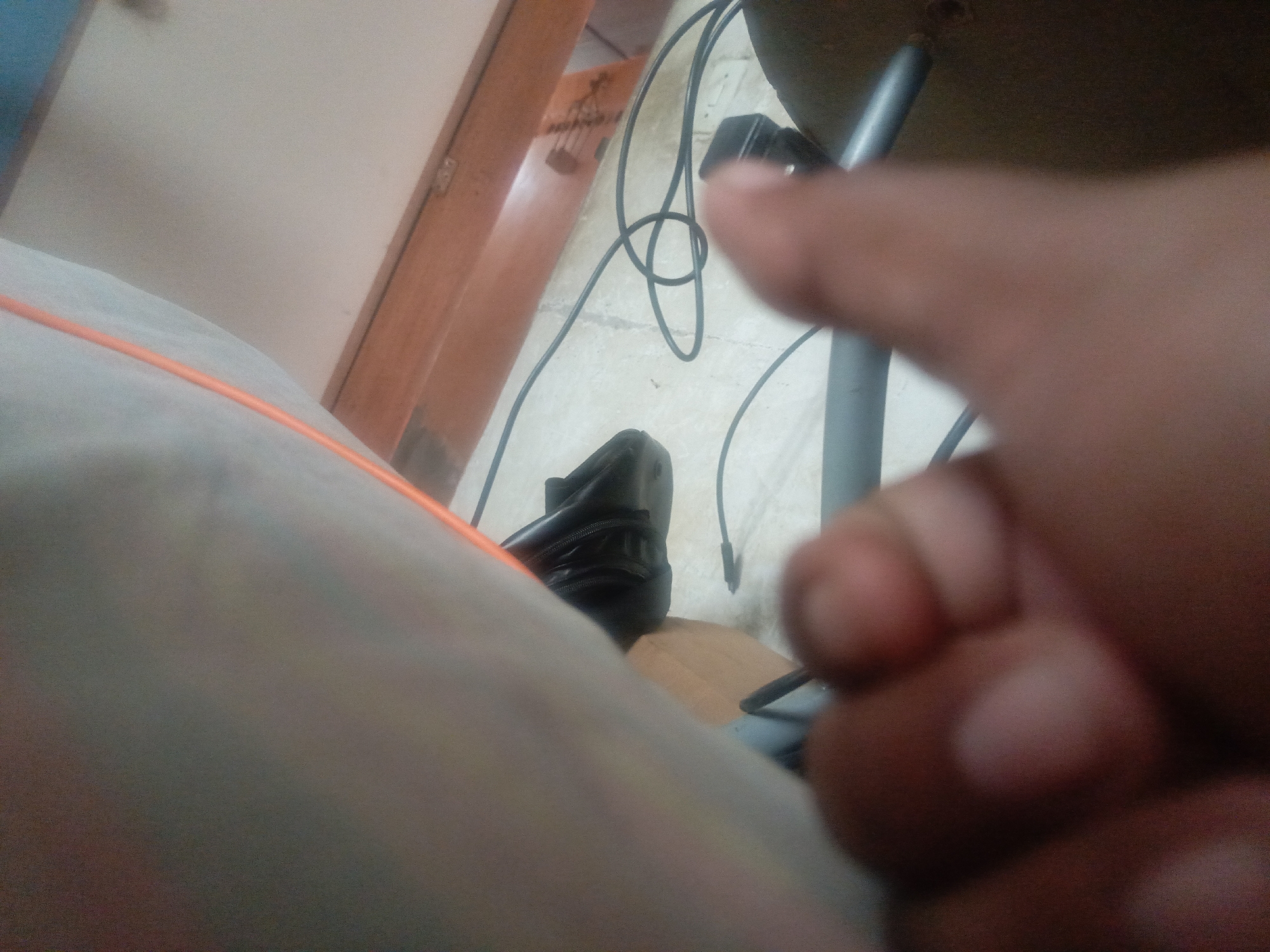 The image shows an indoor setting. A hand is prominent in the foreground, slightly out of focus. A black shoe is visible near a table with gray legs. Wires are tangled on the floor. A portion of a wooden structure is visible in the background, along with a white wall. There is no identifiable information to determine the location is Juan Griego, Venezuela.