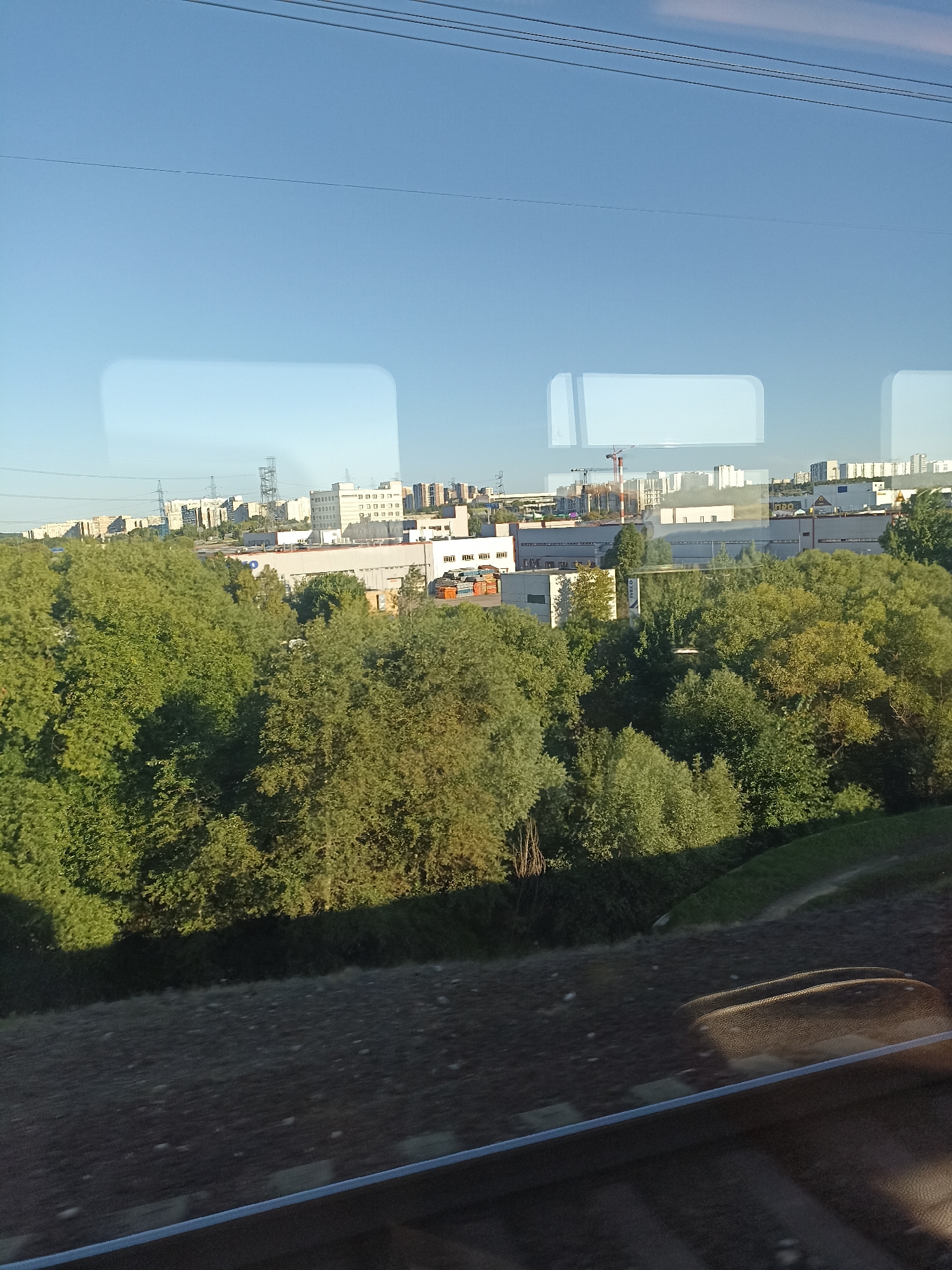 This is an outdoor, daytime, medium-shot photograph taken through a train window, showing a view of a suburban cityscape with a foreground of green trees and a background of buildings. The sky is clear and blue with a few thin wires running horizontally across the top. The train window itself is visible, with two rectangular reflections of the sky in the upper portion of the image. Below the sky and reflections, there is a dense canopy of green trees, predominantly deciduous. Beyond the trees, a cluster of buildings is visible, including low-rise industrial or commercial structures with white and grey facades, some with visible windows and roofing. Taller, multi-story apartment buildings with light-colored facades are visible further in the distance, suggesting a residential area. A construction crane is visible among the buildings on the left side, indicating ongoing development. The bottom portion of the image shows a blurred view of the train tracks and the surrounding embankment, covered with gravel and some vegetation, indicating the train is in motion. The lighting suggests it is either morning or late afternoon, with the sun casting some shadows on the trees and embankment. The scene depicts a transition zone between urban development and natural landscape, typical of the outskirts of a large city like Moscow, Russia.
