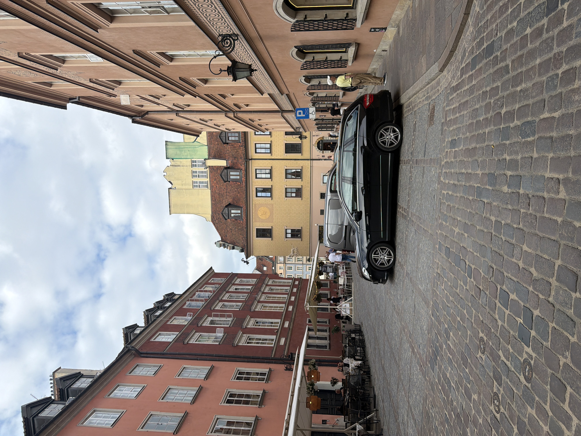 The image captures a street scene in a city with cobblestone paving. On the left, a row of buildings features salmon-pink and dark red facades with multiple windows and mansard roofs. Outdoor seating for a restaurant, complete with tables, chairs, and umbrellas, occupies the sidewalk. Further down the street, a yellow building with a red-tiled roof is visible. On the right, a peach-colored building with arched windows and decorative elements lines the street. A black station wagon is parked on the right side of the street. A man in a light-colored shirt and pants stands near the building on the right, facing away from the camera. Several other people are seen in the background, some walking and others seated at the outdoor restaurant. The sky is partly cloudy.
