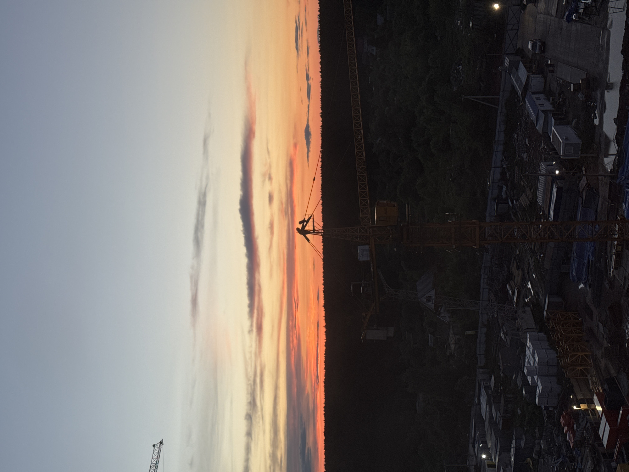 The image shows a construction site in Izhevsk, Russia at sunset. A large yellow crane dominates the scene, standing tall against a sky filled with orange, pink, and grey clouds. To the right of the crane, there's a dark, densely forested area. Below the crane, a construction site is visible, with materials, structures, and equipment scattered around. The sky transitions from light blue on the left to a vibrant sunset near the center. Another crane is visible in the far left, partially obscured. The scene appears to be captured from an elevated vantage point, overlooking the construction site.