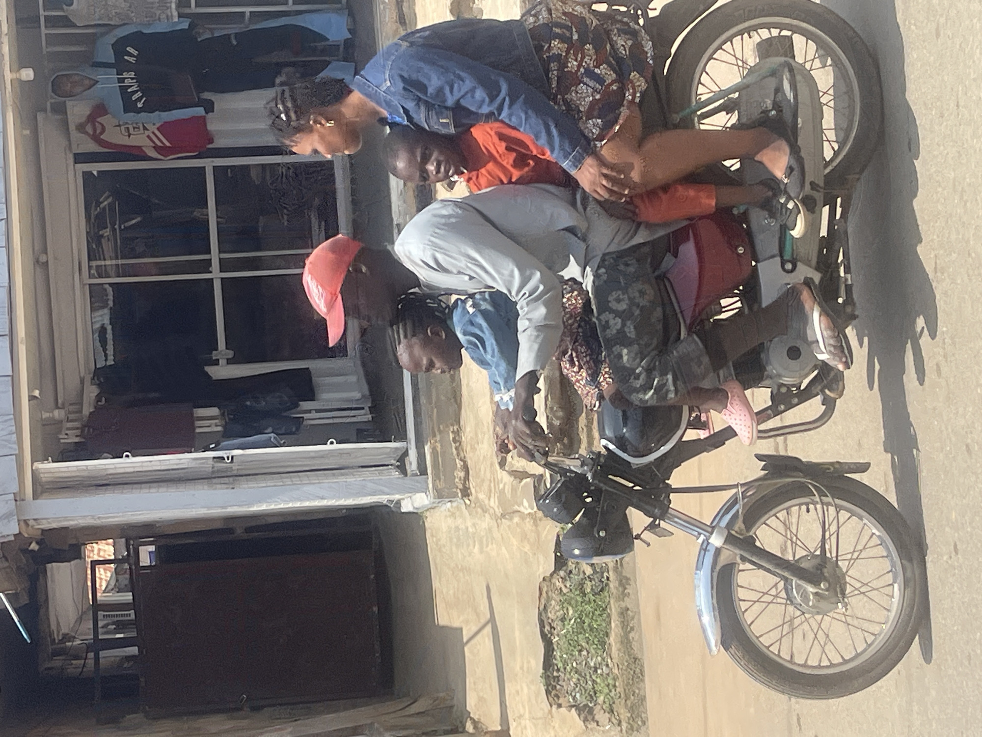 A family of four is riding on a motorcycle in an urban environment, likely in Abuja, Nigeria. The motorcycle is dark-colored with chrome accents, including the front fender and handlebars. A man wearing a light-colored long-sleeved shirt, patterned pants, and a red baseball cap is driving. Seated behind him is a woman wearing a denim jacket and a patterned skirt. Two children are also on the motorcycle; a younger child in an orange shirt is sitting in front of the woman, and a slightly older child in a blue denim jacket is seated in front of the man, between him and the driver. They are riding on a paved road in front of a building with an open storefront displaying clothing on mannequins and racks. The building appears to be a shop or market stall, with a white frame around the storefront entrance. The background shows more of the building and a sunlit, dusty ground area.