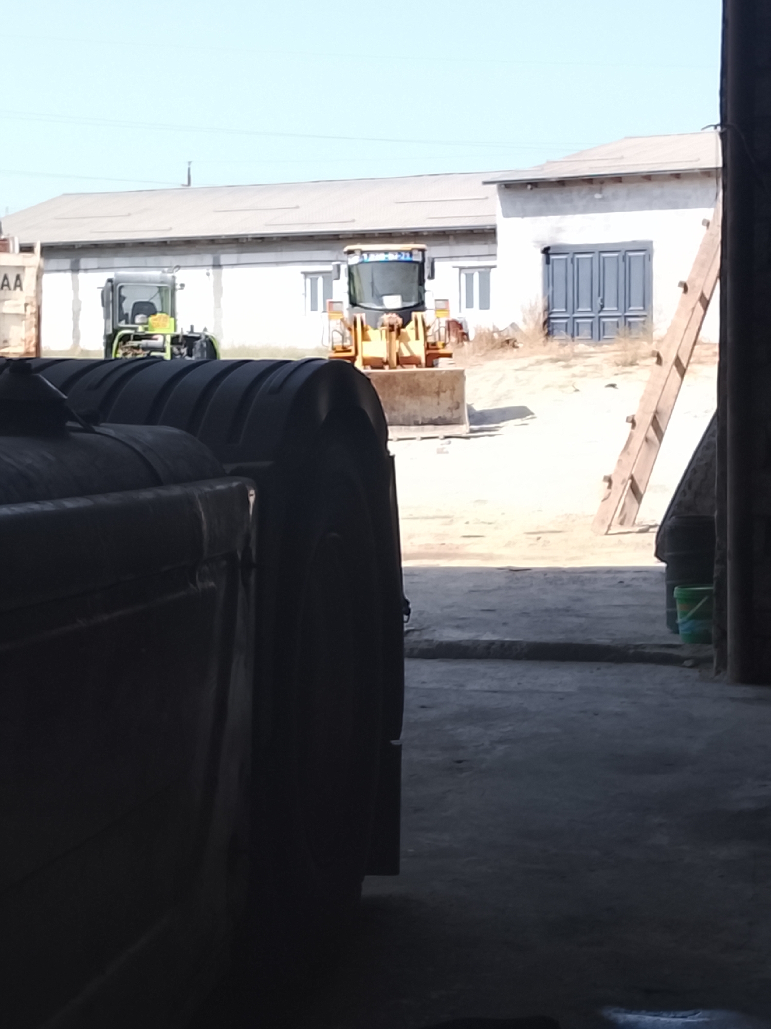 The image depicts an outdoor scene at a facility in Sabir Rakhimov, Uzbekistan. In the foreground, a large, dark, industrial object, possibly a container or piece of machinery, is partially visible on the left side, casting a shadow. Behind it, two yellow construction vehicles are parked on a dirt lot. The vehicle on the left appears to be a tractor or loader with a green cab, while the vehicle on the right is a front-end loader with a raised bucket. In the background, two low-rise buildings with light-colored walls and corrugated metal roofs are visible. One building has large grey double doors. A wooden ladder leans against the right side of the scene, suggesting ongoing work or storage. The sky is clear and blue. The overall impression is of a working yard or storage area for construction equipment.