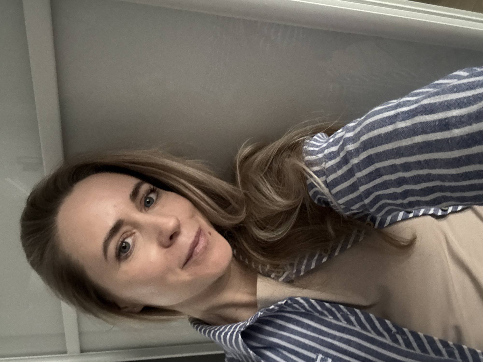 A woman with light brown, shoulder-length hair, blue eyes, and fair skin is taking a selfie. She is wearing a beige shirt under a blue and white striped button-down shirt. The background appears to be a plain, light-colored wall. The location in the city of Chaikovsky, Russia cannot be determined from the image alone.