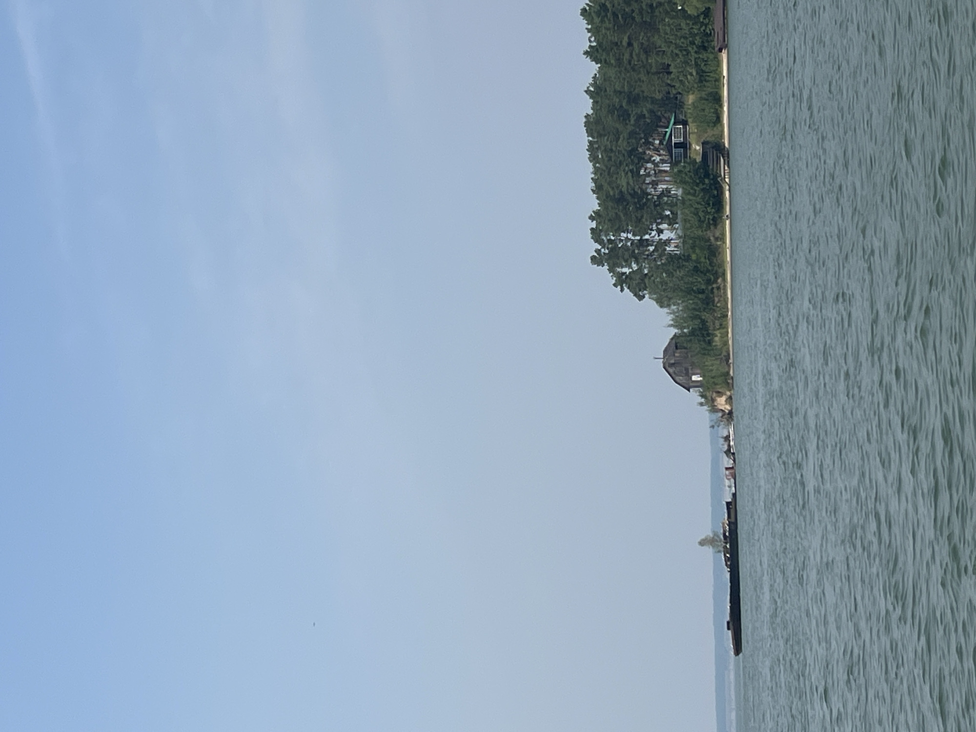 The image shows a wide, calm body of water with ripples under a hazy, pale blue sky. In the distance, a wooded shoreline is visible, with a cluster of trees forming a dense green line. Several structures are nestled within the trees, including a dark-roofed house with a prominent green section and a smaller, older wooden building to its left. A wooden pier with stairs leads down to the water from the house. On the left side of the frame, a long, dark barge sits low in the water, with a small tree growing on it, near some rocky outcroppings. The overall scene suggests a rural or semi-rural setting, likely a lake or a wide river, given the calm water and the presence of the barge. The location is identified as Borovoe, Russia.