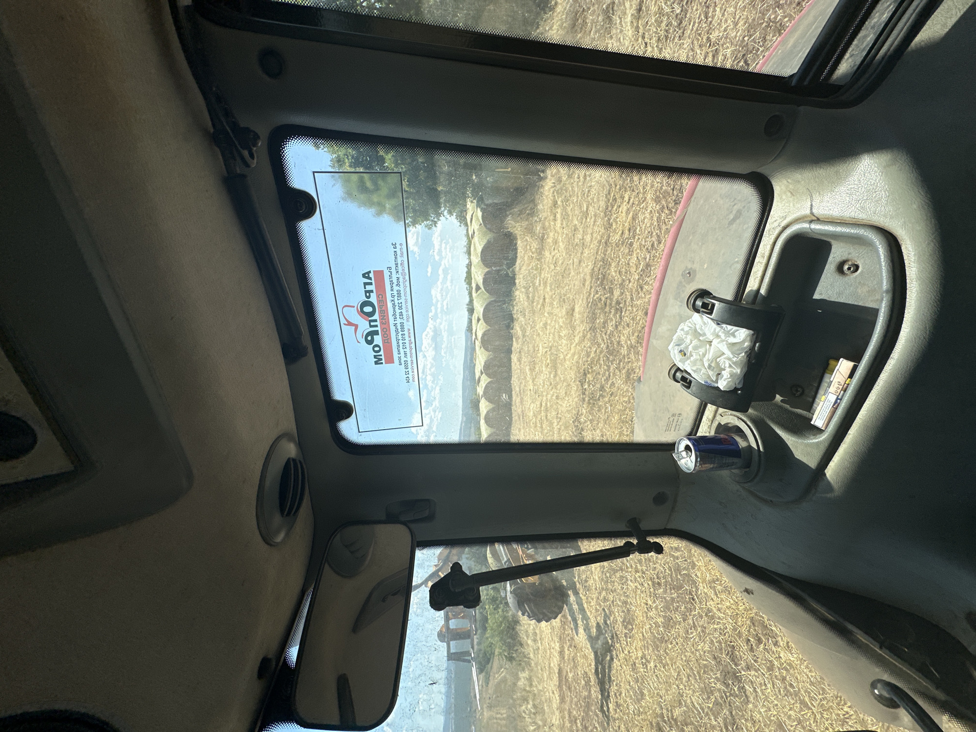 The image shows the interior view from the driver's cabin of a tractor. The cabin is dusty and shows signs of use. A can of Red Bull is placed in a cup holder, next to a crumpled white cloth. Outside the window, a field is visible, covered with hay. Several round bales of hay are lined up in the distance. A large tractor is also visible in the field to the left, and a piece of agricultural machinery is attached to it. The sky is blue with some clouds. A sticker with the logo "Moprorm" is affixed to the windshield. The overall impression is of a farm setting on a sunny day.