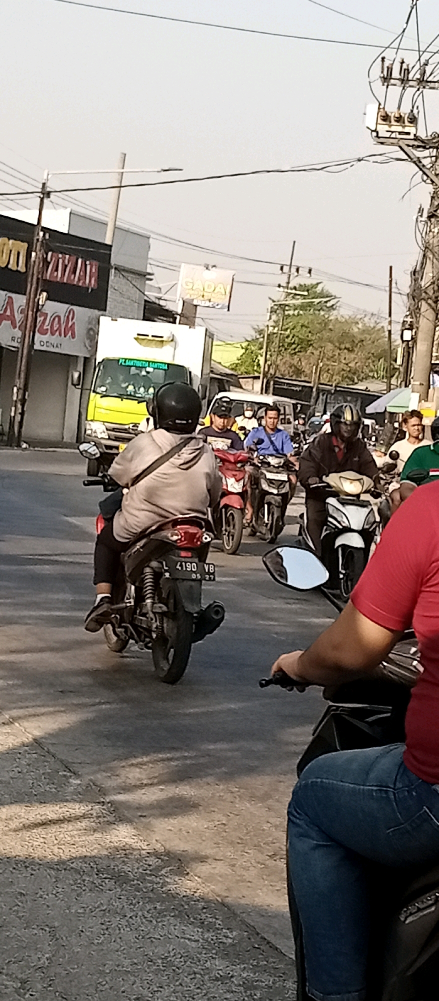 The image depicts a street scene in Kepuh Kiriman, Indonesia, characterized by moderate traffic congestion. Several motorcycles and a yellow truck are visible, indicating a typical urban traffic flow. The environment appears to be a commercial or residential area, with buildings and signs lining the street, including a prominent sign that reads "TIZAR." Power lines and utility poles are overhead, common features in Indonesian streetscapes.
The foreground shows the back of a person on a motorcycle, wearing a grey hooded sweatshirt and a black helmet. The motorcycle's license plate is partially visible, reading "L 4190 YB." To the right, another person on a motorcycle is partially visible, wearing a red shirt and blue jeans.
Further into the scene, several other motorcyclists are present. One individual in a blue shirt is on a motorcycle in the middle ground, alongside other riders in various attire. The yellow truck in the background has text that includes "PT. BAKTI SETIA SENTOSA," suggesting it's a commercial vehicle.
The overall impression is a snapshot of daily life in a bustling Indonesian city, with a focus on the common mode of transportation, motorcycles, and the surrounding urban infrastructure. The lighting suggests it is daytime, with some shadows indicating sunlight.