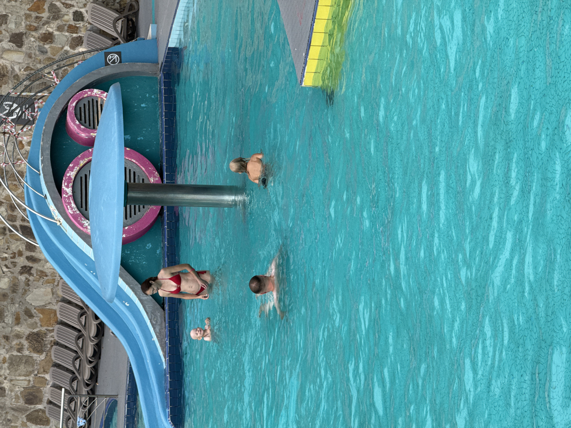 The image depicts a swimming pool area with a blue water slide. In the foreground, four individuals are visible in the turquoise water. A woman in a red bikini stands near the slide, looking down at a small child beside her. Further into the pool, a man is swimming, and a child with blonde hair is facing away from the camera. In the background, rows of lounge chairs are placed on a stone-tiled surface, and the area is bordered by a stone wall. A mushroom-shaped structure with a blue umbrella rises from the pool. The overall scene suggests a public recreational facility. The location is identified as Массандровский поселковый совет, Ukraine.