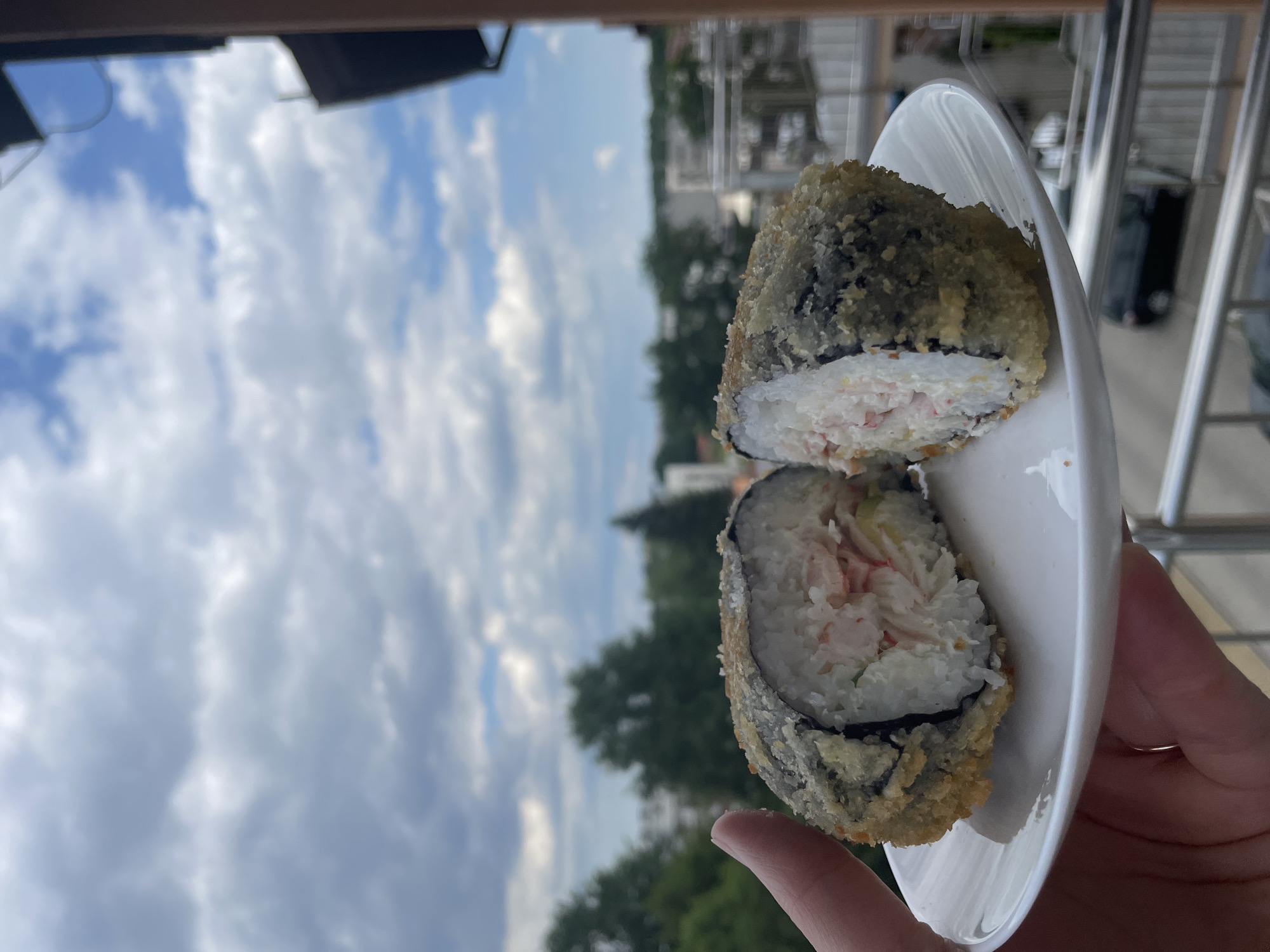 An outdoor, eye-level, medium shot captures a hand holding a white plate with a halved, fried sushi roll. The sushi roll is deep-fried and coated in panko breadcrumbs, revealing a creamy white interior with visible chunks of pink imitation crab meat and some green vegetables. The plate rests on a balcony railing, with a blurred background of trees and buildings under a cloudy sky.