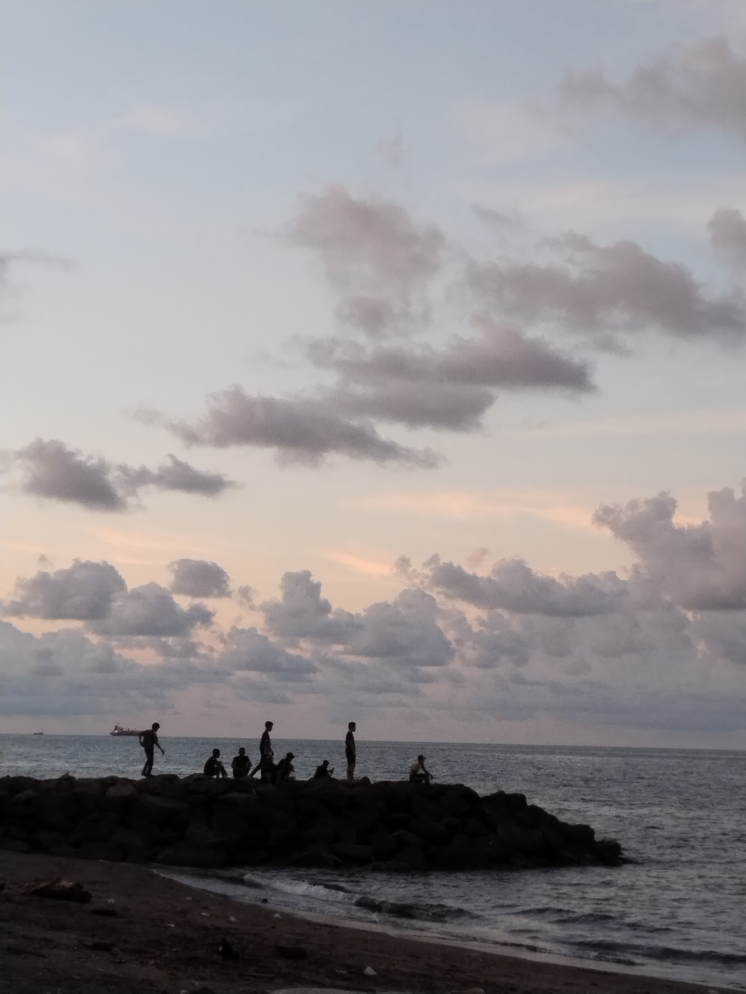 Scene: A group of individuals are on a breakwater at a beach in Lhokseumawe, Indonesia.
People: Seven figures are visible, likely individuals enjoying the beach. They are silhouetted against the sky and the sea. One appears to be standing while another is sitting.
Objects: The prominent feature is a breakwater. In the distance is an ocean, under an open sky with clouds.
Environment: The sky is filled with clouds, indicating possibly dusk. A beach leads to the water, with waves breaking near the breakwater.