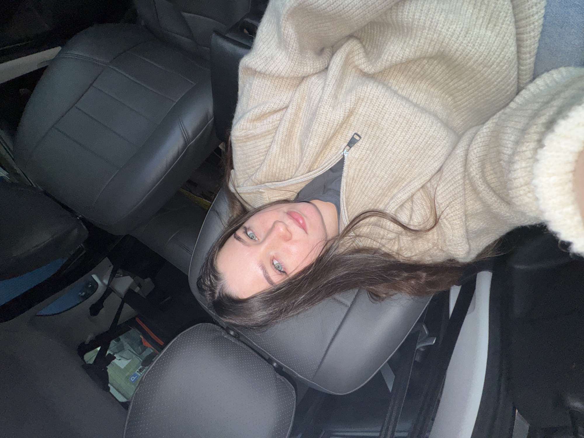 A young woman is depicted lying down inside what appears to be a car or similar vehicle. She has long, dark brown hair, blue-green eyes, and light pink lips, looking directly at the camera with a neutral expression. She is wearing a light beige or cream-colored chunky knit cardigan or zip-up sweater over a darker shirt, with a hint of blue denim fabric visible near her right shoulder.
Her head rests on a dark gray or black car seat, which features distinct stitching and some perforated textures, suggesting a leather or synthetic leather material. Other car seats and interior components, such as door panels and the vehicle's floor, are visible in the background, appearing dark and slightly indistinct due to low lighting. The angle of the shot suggests she is lying across the seats, possibly posing for a selfie. The overall setting is indoors or at night, given the muted lighting conditions. There is no visible text in the image. The event taking place is a posed photograph within a vehicle.