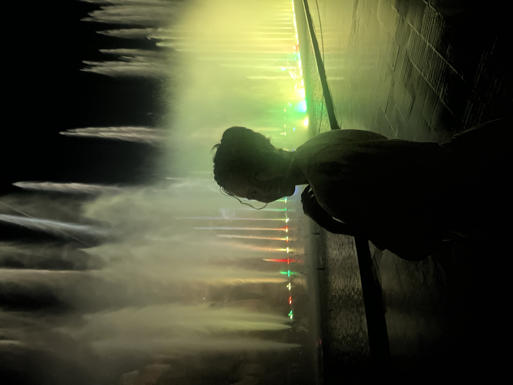 At night, a person with dark hair observes a colorfully lit fountain display, possibly in the city of Nha Trang, Vietnam. The person's silhouette suggests they are leaning over a wall, gazing down at the fountain. The fountain has multiple streams of water illuminated with various colors, including yellow, green, red, and white. The background is dark, but some faint clouds are visible in the upper-left corner. The right side of the image includes a wall.