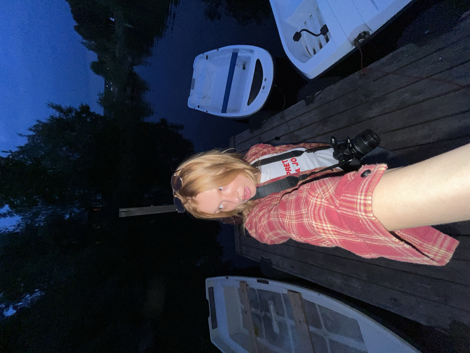 A young woman with blonde hair, wearing a red and yellow plaid shirt over a white t-shirt, stands on a wooden dock at dusk. She is holding a camera and taking a selfie, looking towards the viewer with a surprised expression. Her sunglasses are pushed up onto her head. To her left is a white rowboat, and to her right, a larger white boat floats on dark water. The background shows trees and a dark blue sky. This scene is likely located in the city of Chaikovsky, Russia, given the presence of boats and a dock, common features along the Kama River, which flows through the region.