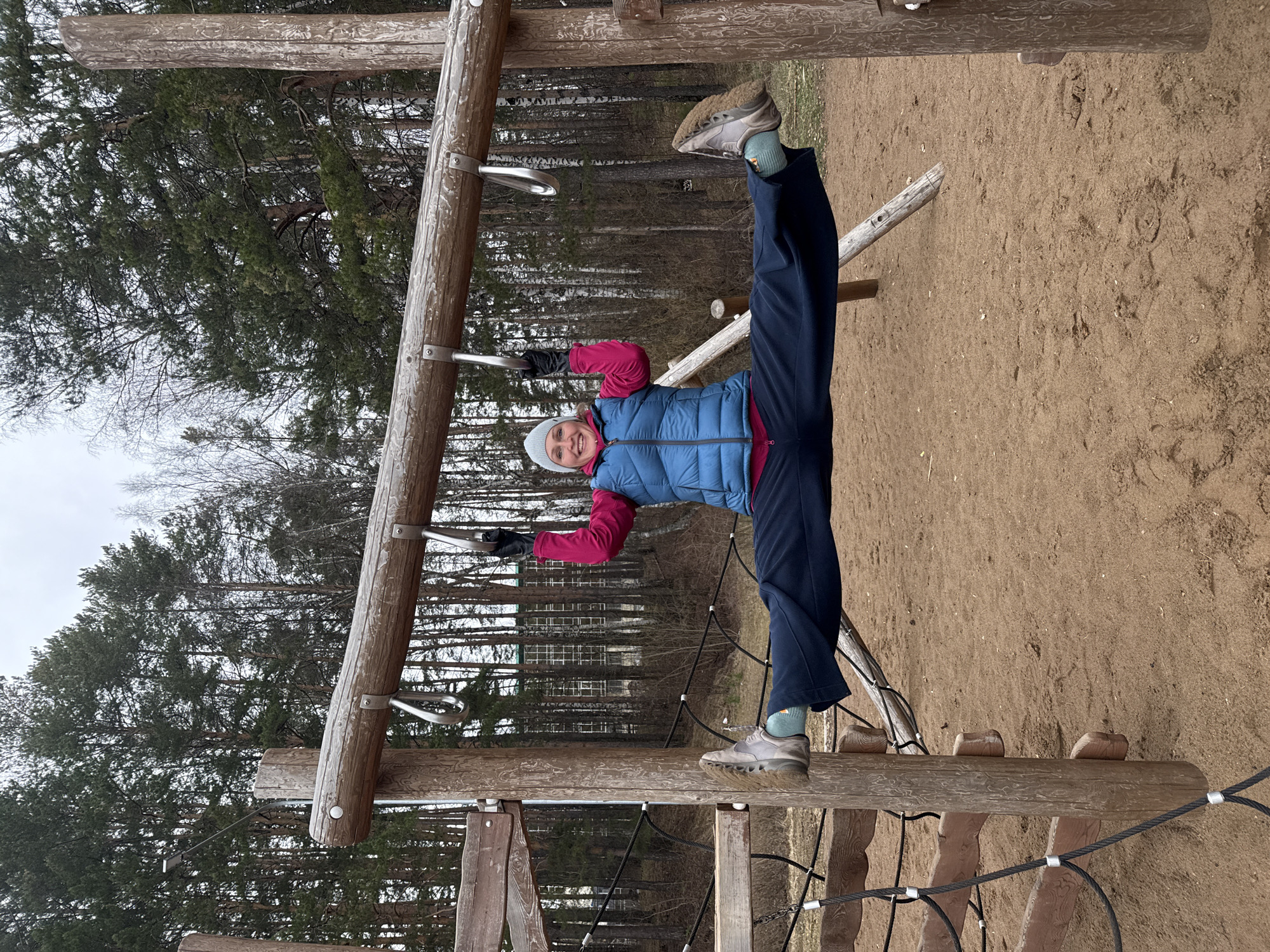 An adult is captured in a playful and athletic pose, hanging upside down from wooden monkey bars in an outdoor setting. She is performing a split, with her hands gripping the horizontal bars and her legs extended wide; one foot rests on a lower wooden beam of the playground structure. She is smiling broadly at the camera.
The person is dressed for cool weather, wearing a blue puffer vest over a pink long-sleeved top, dark blue athletic pants, a grey beanie, and grey athletic shoes with light blue socks. Black gloves are also visible on her hands.
The scene takes place on what appears to be a playground or outdoor fitness area, characterized by robust wooden structures. The ground is sandy or dirt-covered. In the background, there is a dense forest primarily composed of tall evergreen trees, along with some bare deciduous trees, suggesting an early spring or late autumn season in Chaikovsky, Russia. The sky is overcast, indicating a daytime shot without direct sunlight. Through the trees in the distance, a multi-story building with numerous windows can be faintly seen, hinting at an urban or suburban edge to the forested area. No visible text is present in the image.