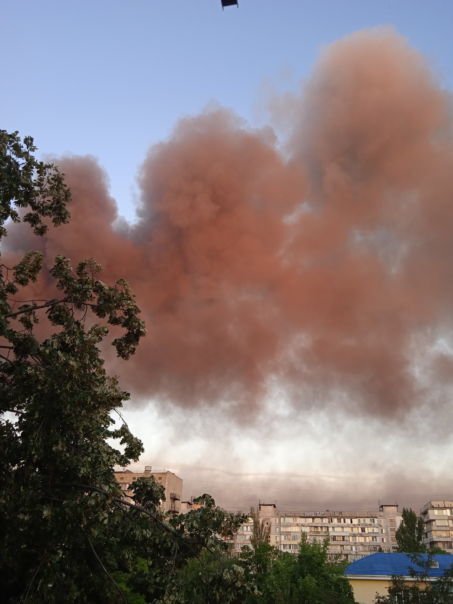 A vast plume of thick, reddish-brown smoke billows across a pale blue sky, obscuring the upper portions of several apartment buildings. The smoke appears to be emanating from a fire or industrial source, casting a warm glow on its edges. In the foreground, a large tree with dark green leaves partially obstructs the view of the buildings. The buildings themselves are typical of Soviet-era construction, with multiple floors and rows of windows. Some have visible external staircases or air conditioning units. The overall scene suggests an urban environment experiencing an event that is producing significant smoke.