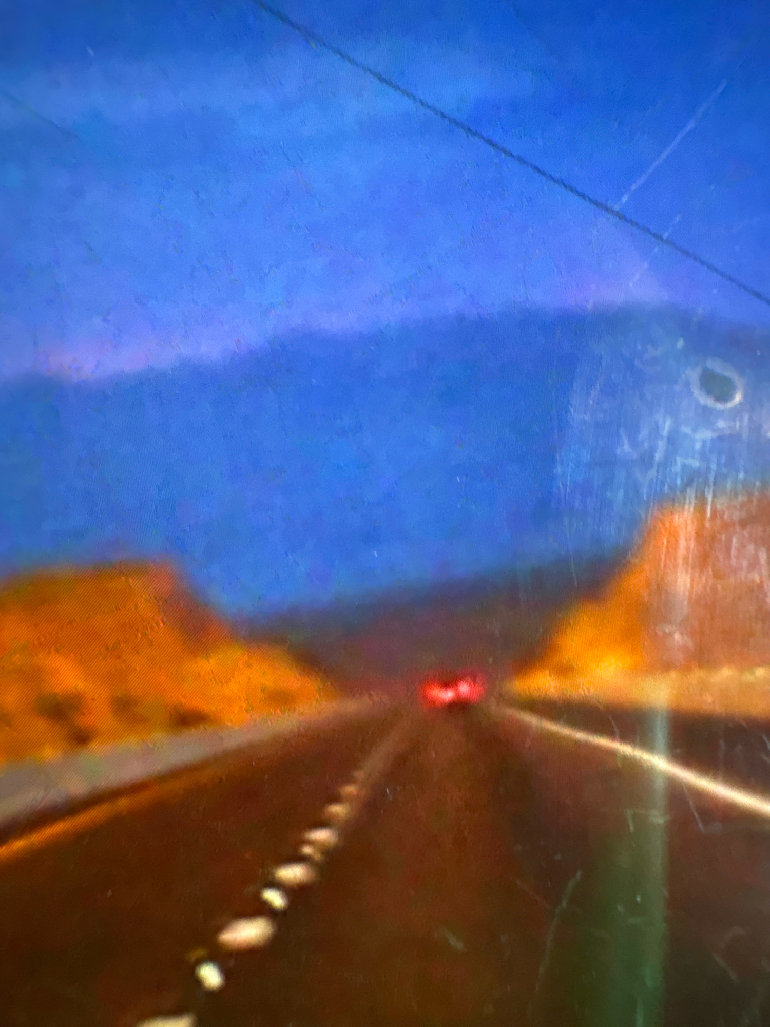 The image depicts a road with a car in the distance. The sky is a deep blue, and there are hills or mountains in the background. The road is dark and appears to be wet, with white lines marking the lanes. There are some orange and yellow areas on either side of the road, which could be foliage or rock formations. A thin black line runs diagonally across the top of the image, possibly a wire or cable. The image is out of focus and appears to be a painting or a heavily filtered photograph, with visible scratches or texture on the surface. There are no people or specific landmarks to identify a particular location in Vagator, India.