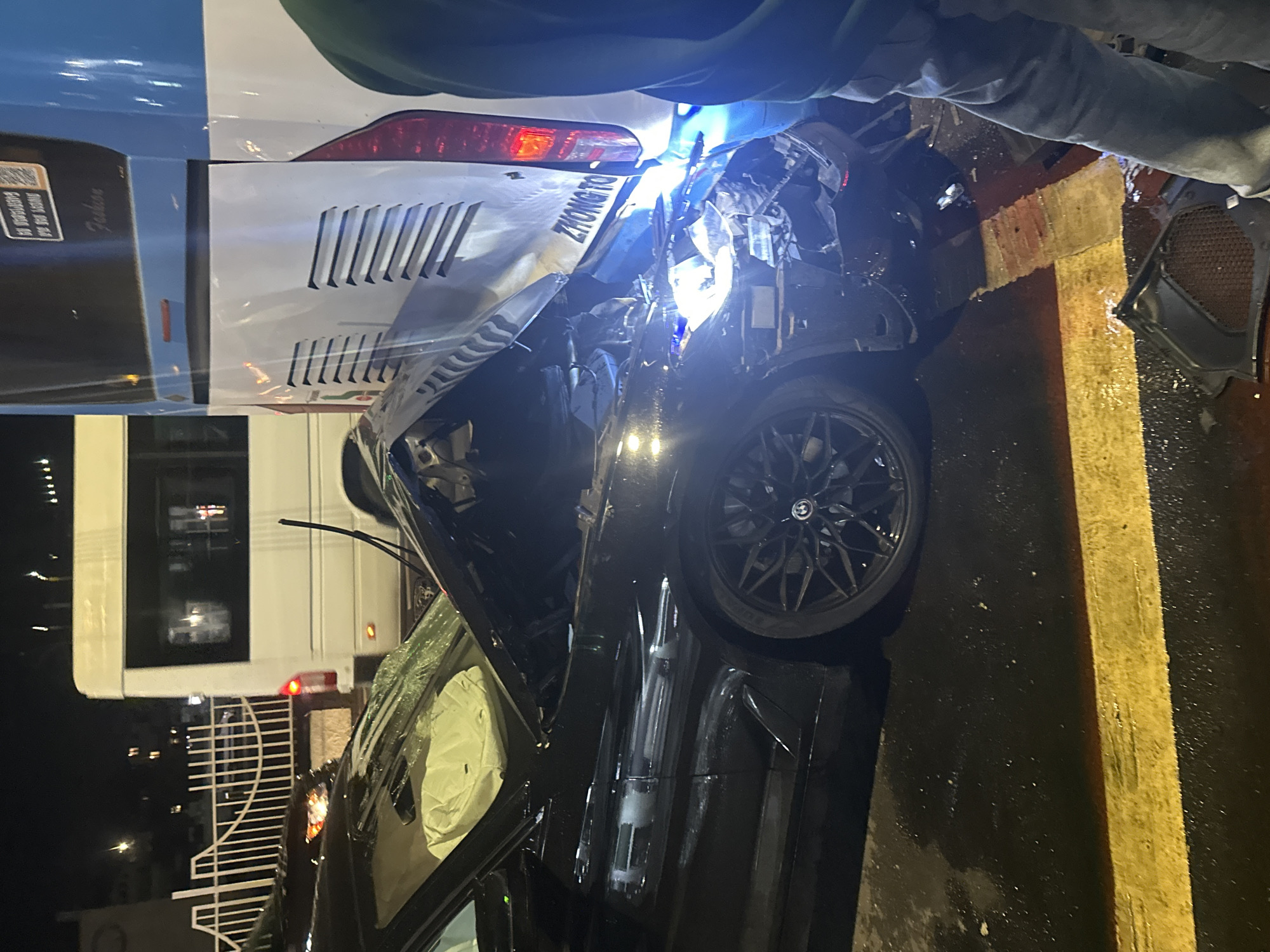 The image shows the aftermath of a car accident at night. A black vehicle, possibly a BMW judging by the wheel's logo, is heavily damaged. Its front end is crushed, the windshield is cracked, and the airbags have deployed. The car appears to have collided with the rear of a white van or bus, which shows some damage as well. The van has the word "ZHONGTO" on the back. Debris is scattered around the black car. The scene is on a street with a yellow lane marking. Based on the evidence found on the rear of the white van or bus, its possible location is Yerevan, Armenia. A person in casual clothing is standing near the vehicles, possibly observing or inspecting the damage. A gate is visible in the background.