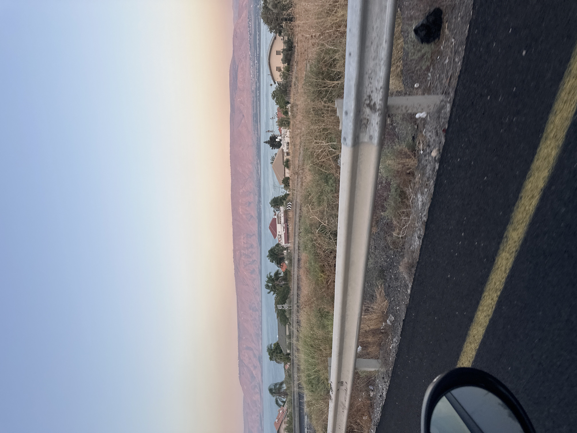 The image captures a scenic view of the Sea of Galilee from a roadside in the Emek HaYarden Regional Council, Israel. The sky transitions from a pale blue at the top to a soft, peachy hue near the horizon, suggesting either sunrise or sunset. A vast body of water, the Sea of Galilee, dominates the middle ground, with rolling hills in shades of pink and mauve forming the distant landscape.
In the foreground, a grey metal guardrail runs horizontally across the image, with dry, brown vegetation and some scattered debris visible behind it. The road surface, a dark asphalt with a single yellow line, occupies the lower portion of the frame, and a side-view mirror of a vehicle is partially visible on the left, indicating the perspective is from inside a car.
A cluster of houses with red and tile roofs, interspersed with palm trees and other greenery, are situated along the shoreline. The architecture appears to be Mediterranean-style. The overall impression is one of a tranquil, natural landscape with residential development. The lighting suggests a serene, possibly early morning or late evening atmosphere.