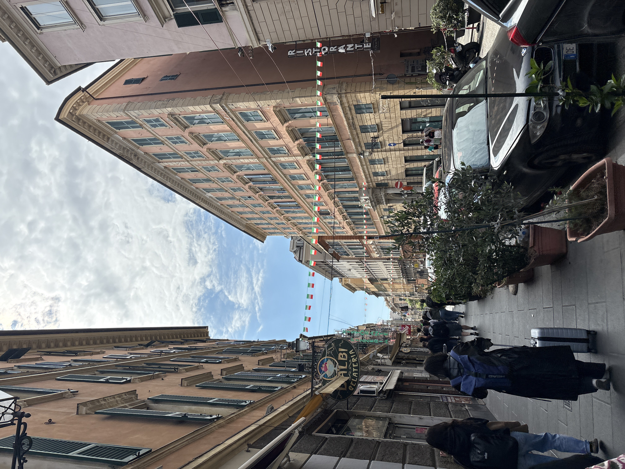 The image captures a street scene in Genoa, Italy. Tall buildings line the narrow street, their facades displaying multiple windows with shutters. Italian flags are strung across the street. A restaurant sign is visible. On the right side of the frame, a dark-colored car is parked alongside the sidewalk, where several potted plants are placed. Pedestrians are walking along the sidewalk, some pulling luggage. The sky is partially visible between the buildings, showing a mix of blue and white clouds.