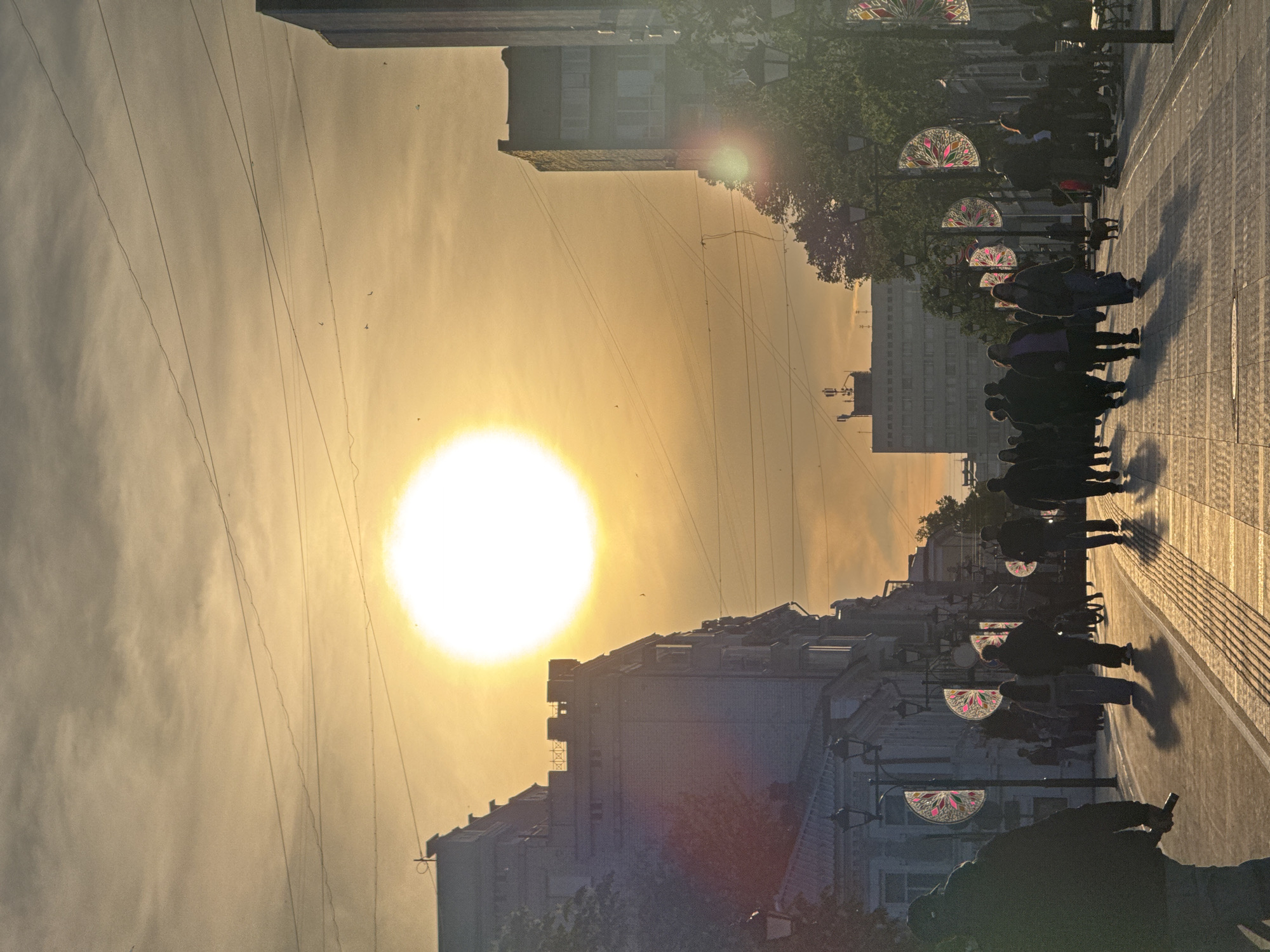 The image depicts a street in Saratov, Russia, at sunset. The sun is setting directly down the street, casting a strong, warm light. Silhouetted people are walking along the sidewalk, their shadows stretching long. The street is lined with buildings, also silhouetted, and decorative streetlights with stained-glass fan patterns. Overhead, electrical wires crisscross the sky.
