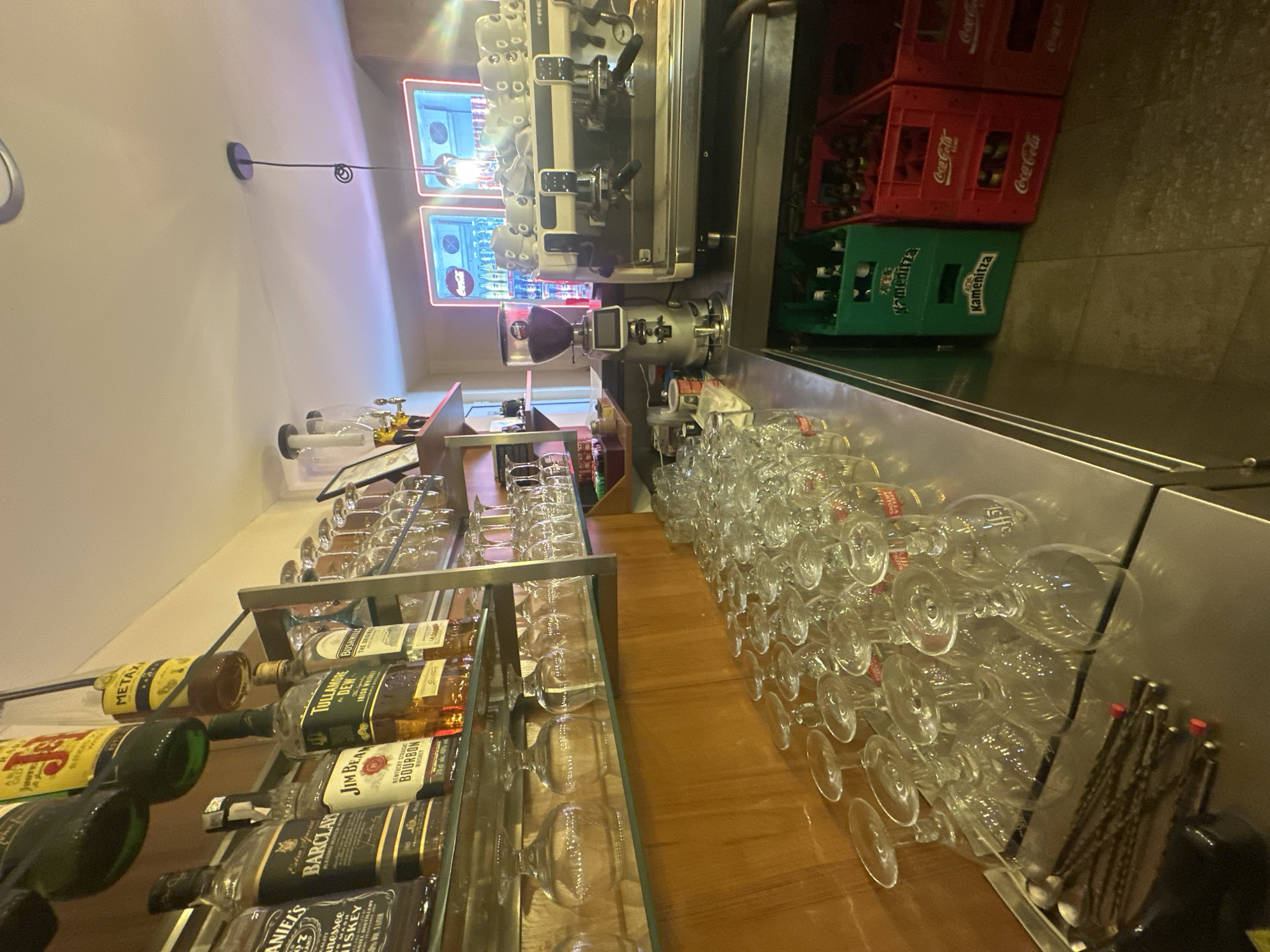 The image, captured in a vertical orientation, depicts a well-equipped bar or service counter, likely within a cafe or restaurant in Lom, Bulgaria. The main subjects are various alcoholic beverage bottles, numerous drinking glasses, professional bar equipment, and beverage crates.
In the foreground, a polished wooden counter stretches horizontally, covered with an extensive array of clear glass stemmed and regular glasses, many inverted and stacked, suggesting they are clean and ready for use. Some glasses feature brand logos like "STELLA ARTOIS" and "LEFFE." To the left, a multi-tiered glass shelf unit holds a variety of liquor bottles. Visible brands include "METAXA," "BARCLAY BLENDED SCOTCH WHISKY," "JIM BEAM KENTUCKY STRAIGHT BOURBON WHISKEY," "TULLAMORE DEW IRISH WHISKEY," "BUSHILLS THE ORIGINAL IRISH WHISKEY," and "Jack Daniel's Tennessee Whiskey" (specifically "Tennessee Whiskey").
Further back on the counter, a large, modern espresso machine with multiple group heads and a partially visible "PREMIUM" label is prominent, alongside a coffee grinder. Behind this equipment, two rectangular, neon-lit display panels, possibly advertising, show the "Coca-Cola" logo and bottled beverages. On a lower level to the right of the counter, several plastic beverage crates are stacked; a green crate clearly displays "Kamenitza," a Bulgarian beer brand, while two red crates are marked with the "Coca-Cola" logo.
The scene is illuminated by overhead artificial lighting and the glowing displays, indicating an indoor setting. No people are present, and no direct activity is taking place, but the organized arrangement of items suggests a functional and active beverage service area.