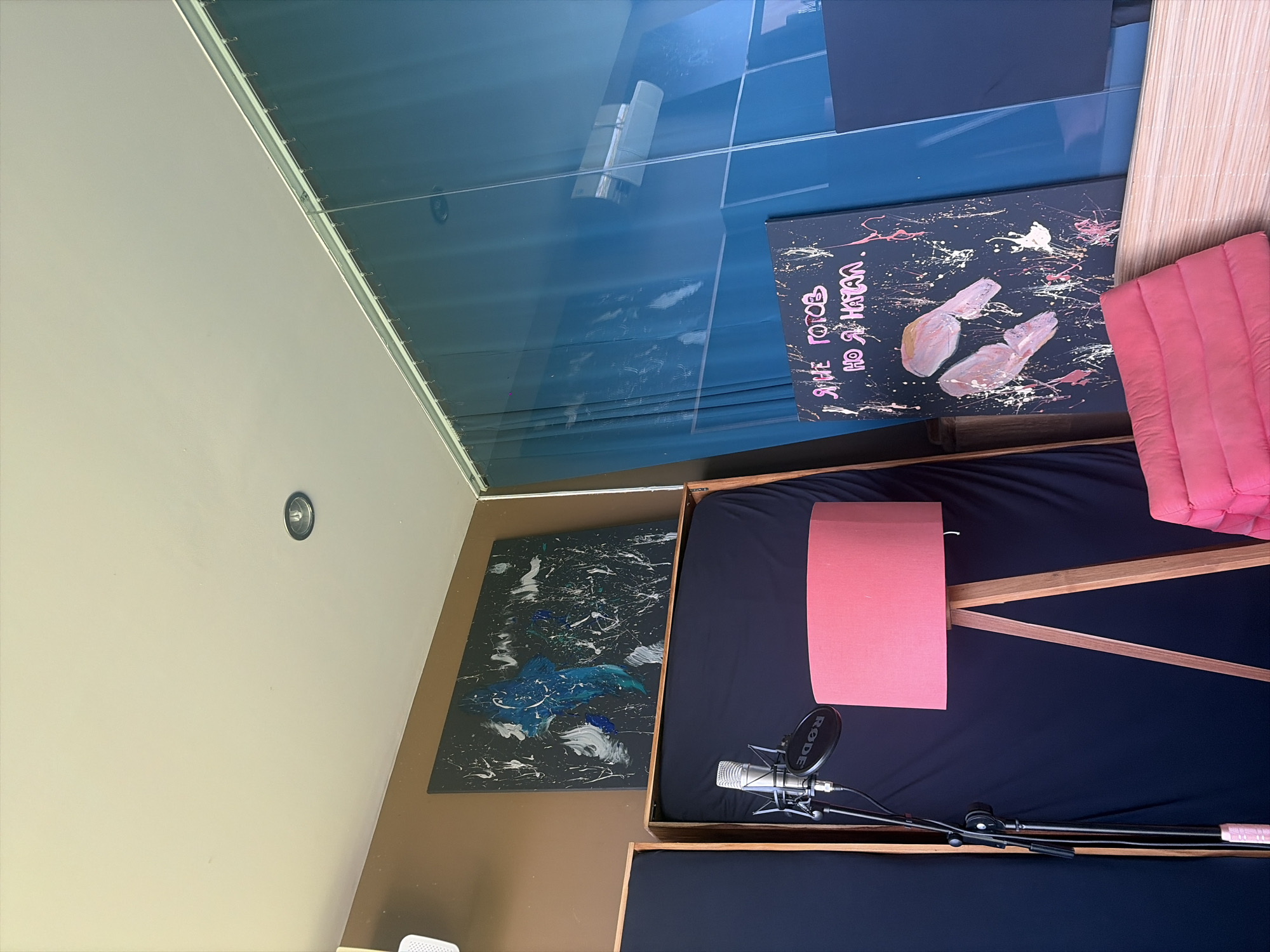 The image shows an indoor scene, likely a room set up for recording or artistic creation. In the foreground, a pink cushioned seating area is visible, next to a floor lamp with a pink shade and a wooden tripod base. To the left of the lamp, a microphone on a stand is positioned in front of a dark fabric backdrop. Above the backdrop, a large abstract painting with black, blue, and white elements is mounted on a brown wall. Further to the right, another abstract painting with pink and white splatters and what appear to be stylized handprints is displayed. This painting also features text in pink lettering. The background is dominated by a translucent blue curtain or screen, behind which various elements are faintly visible, including what looks like an air conditioning unit. The ceiling has a recessed light fixture. The overall impression is of a creative space, possibly a home studio. The location in Mengwi, Indonesia, is not evident from the visual information in the image.
