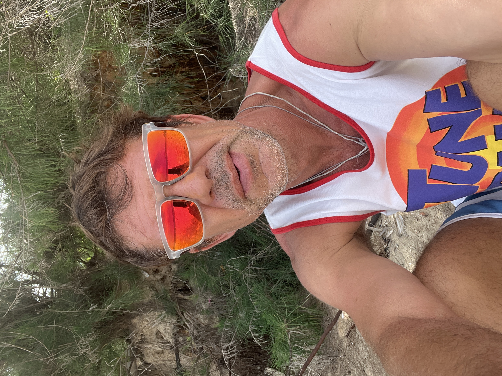 A man with brown hair, sunglasses with red lenses, and a white tank top with red trim is sitting in front of some bushes on a sandy area. The man is looking at the camera and smiling. He is wearing a silver necklace. The tank top has a "Tune Squad" logo on it. The man is in Mui Ne, Vietnam.