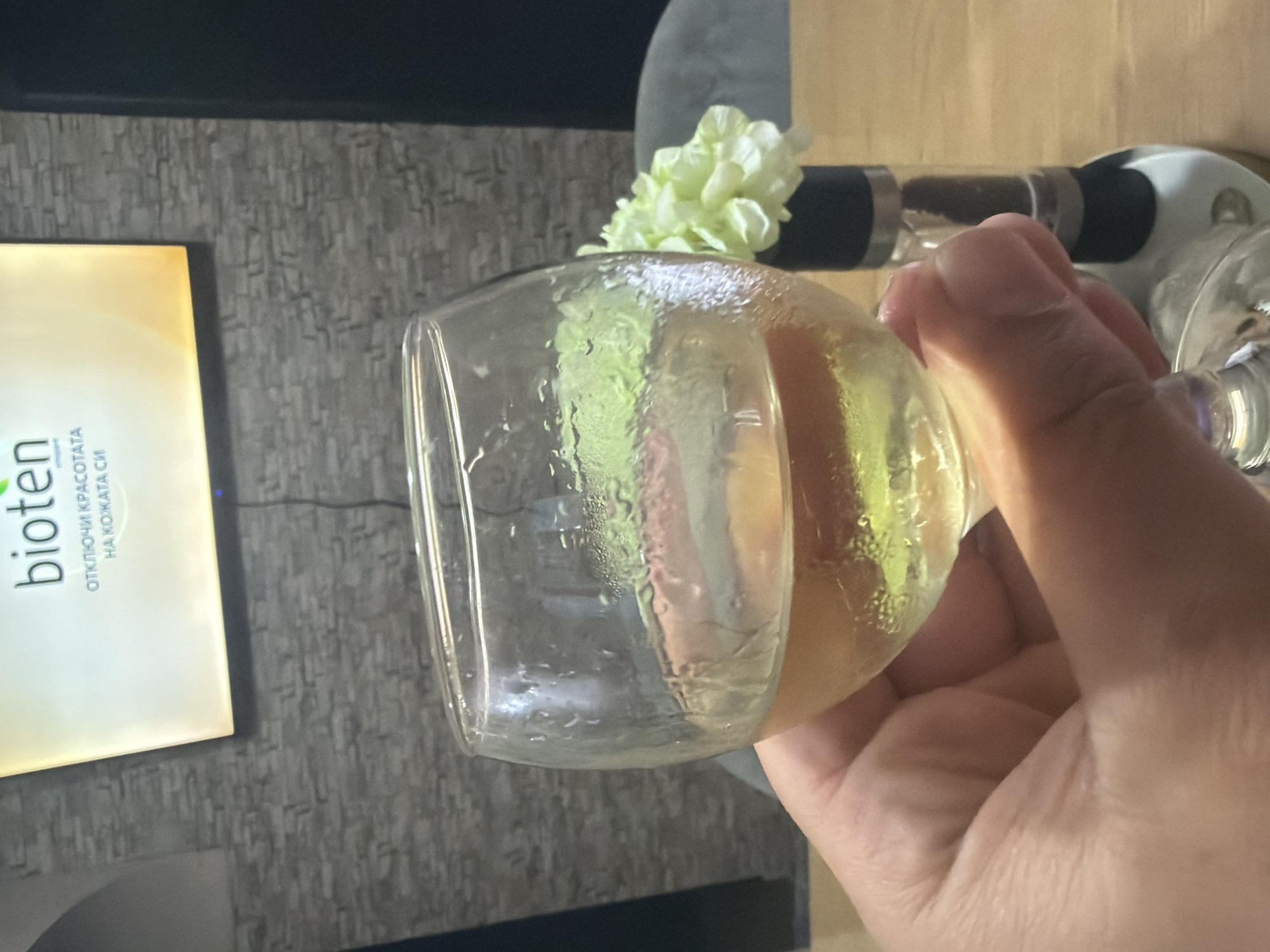 The image displays an indoor scene, likely in Shumen, Bulgaria, featuring a person's hand holding a glass. The hand, with light skin, grips a clear glass filled with a pale yellow liquid, suggesting white wine or a similar beverage. Condensation droplets are visible on the exterior of the glass, indicating the drink is cold.
In the background, a partially visible bottle with a black stopper is present, possibly an alcoholic beverage bottle. A small cluster of light green or white flowers acts as a decorative element behind the glass and bottle. To the left, a digital screen or tablet displays advertising content. The brand name "bioten" is clearly visible, followed by Cyrillic text: "ОТКЛЮЧИ КРАСОТАТА НА КОЖАТА СИ," which translates to "UNLOCK THE BEAUTY OF YOUR SKIN." The background features a textured wall, possibly with a stone or abstract pattern. The overall impression is one of a relaxed setting, perhaps during an evening or leisure time.