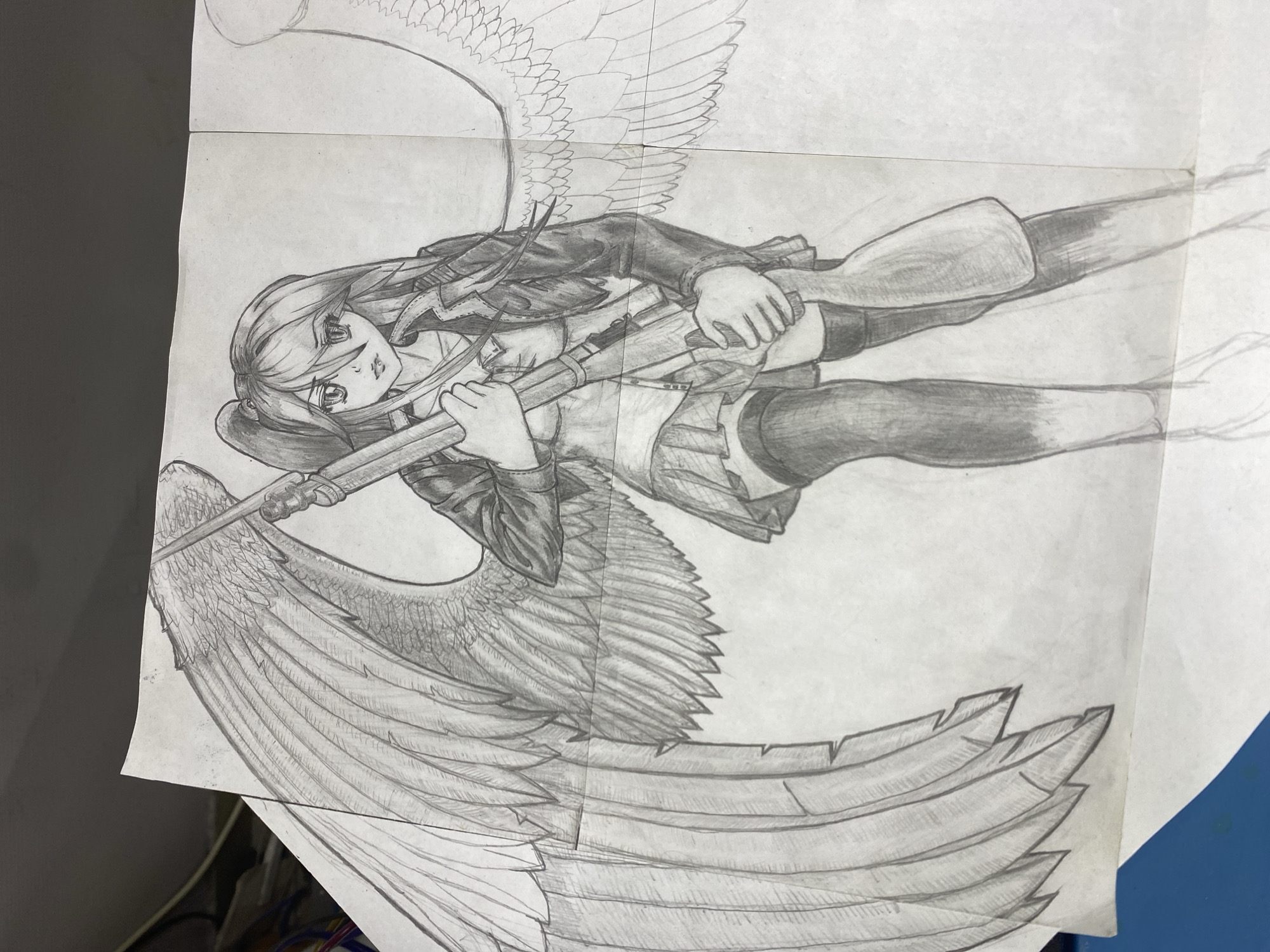 The image is a pencil drawing of an anime-style female character with wings, holding a rifle. She is wearing a jacket, a skirt, and thigh-high socks. The wings are detailed and large. The drawing appears to be on two separate pieces of paper that are taped together. The overall aesthetic suggests a combination of fantasy and military themes. There is no indication of the city of Chaikovsky, Russia in the image. The drawing is on a white background, possibly a table or desk. Cables can be seen in the bottom left corner.