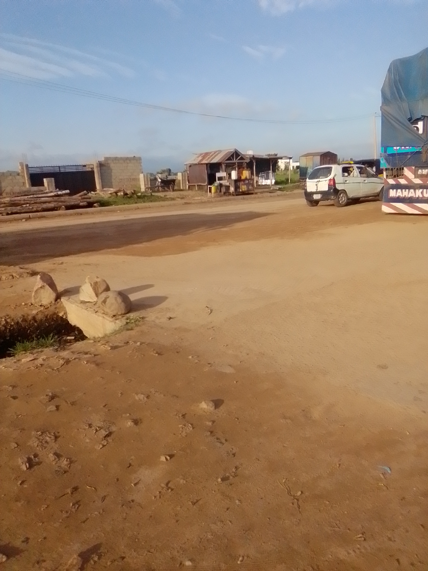 The image depicts an outdoor scene in Kwadon, Nigeria, under a clear blue sky with wispy clouds. The foreground is dominated by a dirt road and rough terrain with scattered rocks and small plants. In the midground, several structures are visible, including a building with a corrugated metal roof, a fence, and piles of lumber. A white car is parked on the right side of the road, next to a large truck covered with a blue tarp. The truck has "MAHAKU" written on its side. There are no people clearly visible in the image. The overall impression is one of a dusty, somewhat undeveloped area, possibly a construction site or a roadside market.